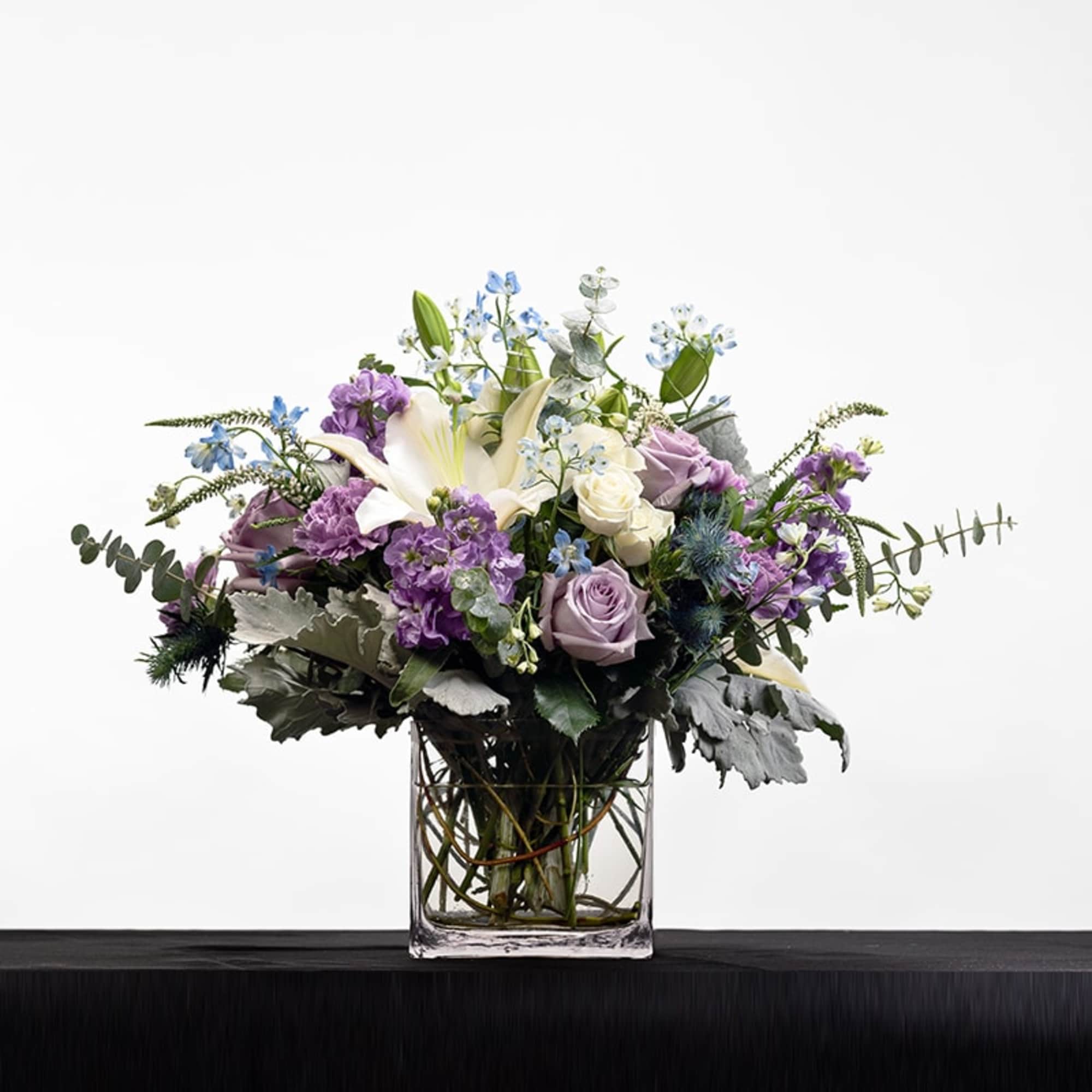 Lavender and white flower arrangement with lilies and roses in a clear rectangular glass vase.