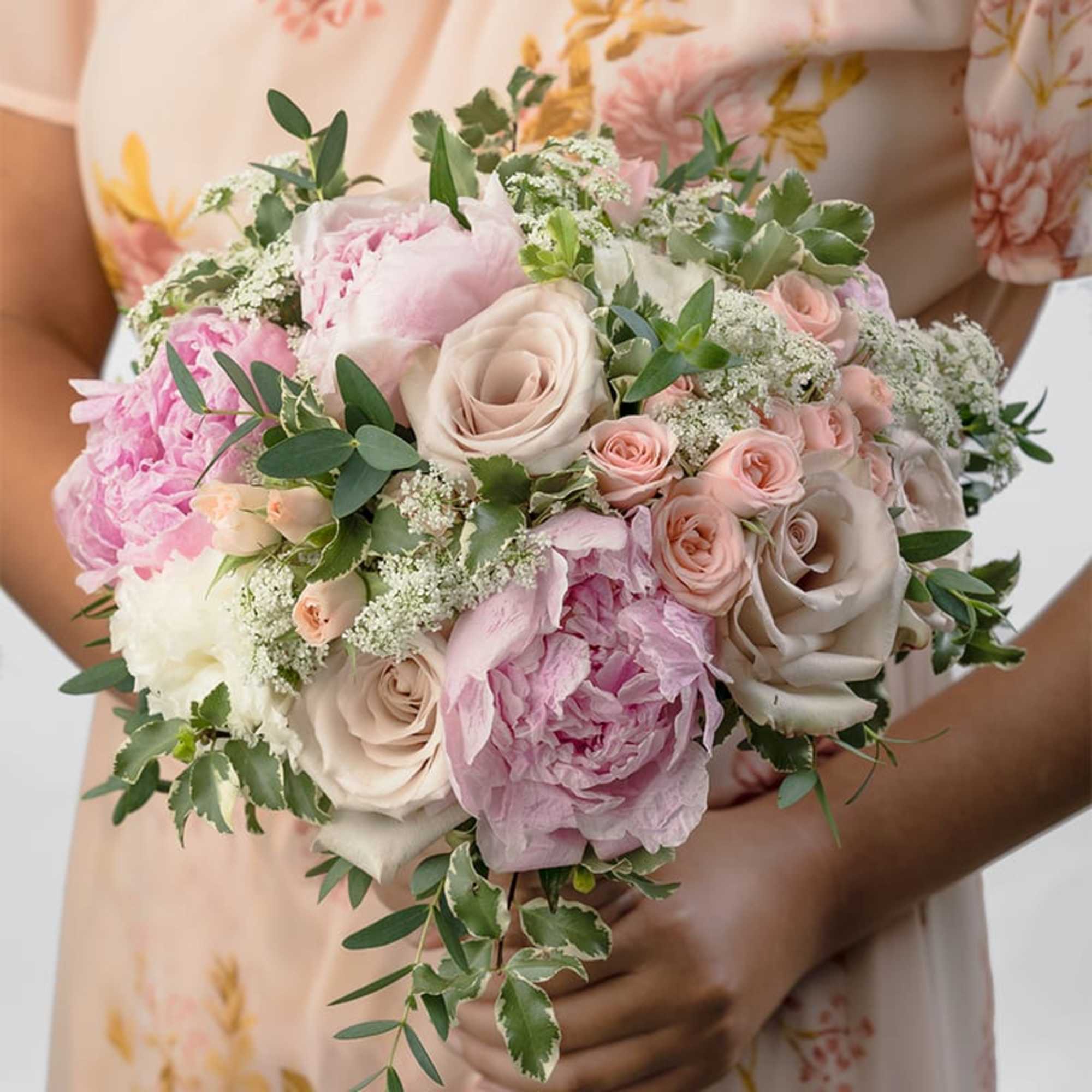 Hand-tied bouquet of blush roses, pink peonies, and white filler flowers held by a person in a floral dress