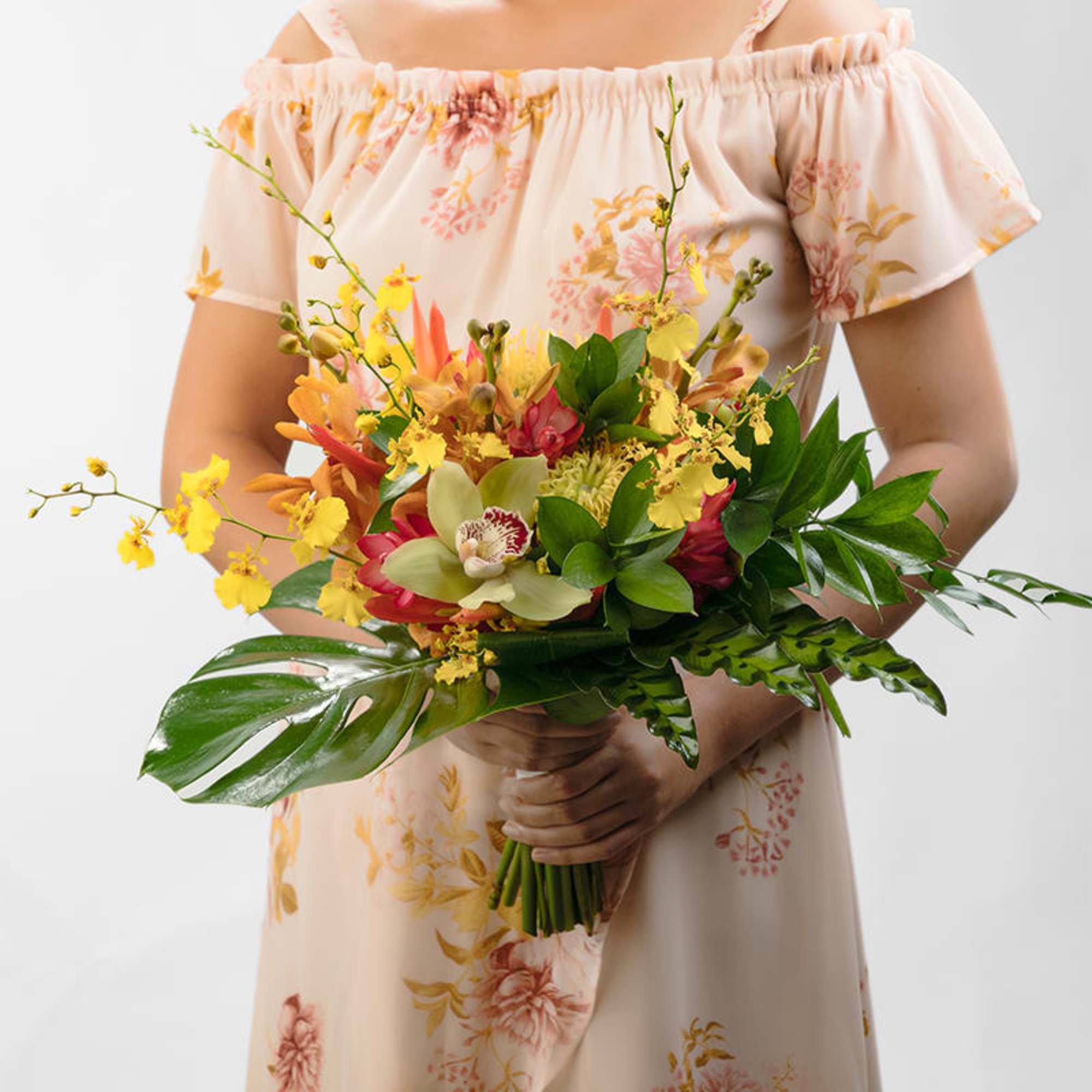 Person in a floral dress holding a tropical bouquet with yellow orchids and red and green flowers.