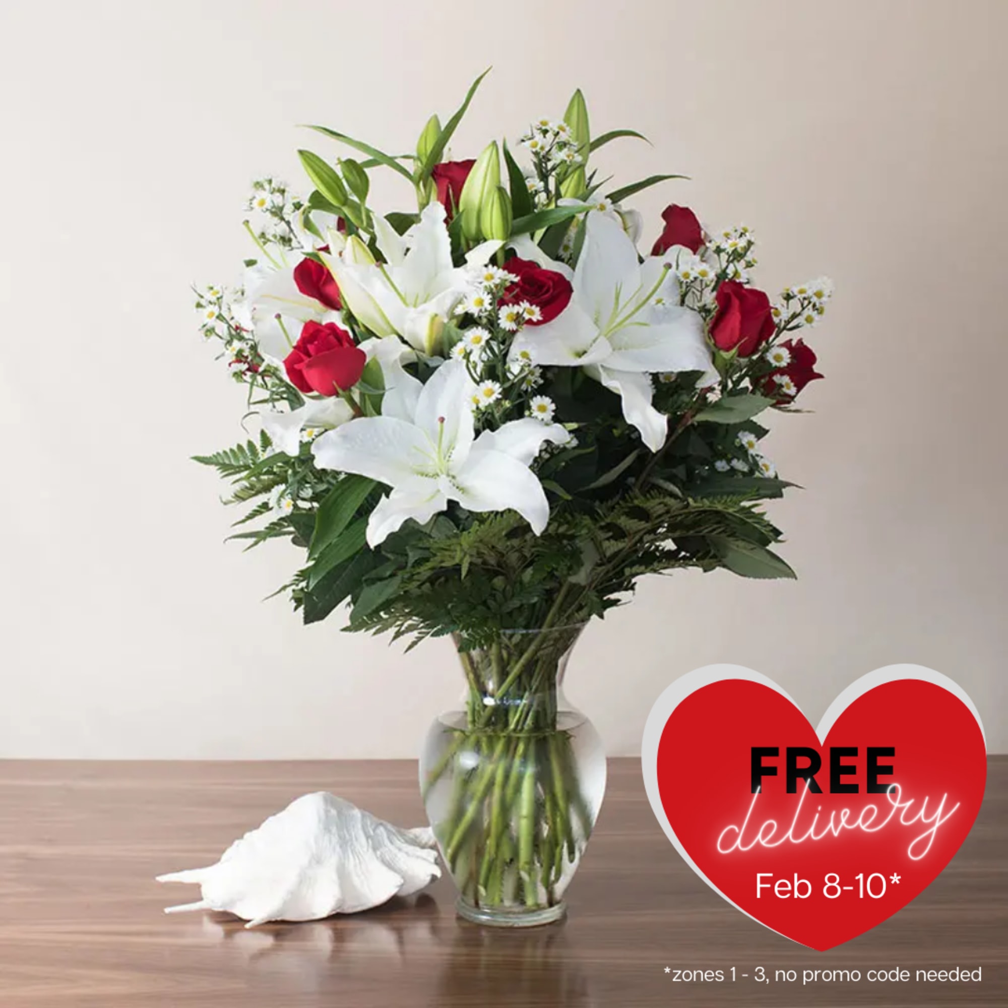 Clear glass vase of white lilies, red roses, and small white blooms on a wooden table with a white seashell.