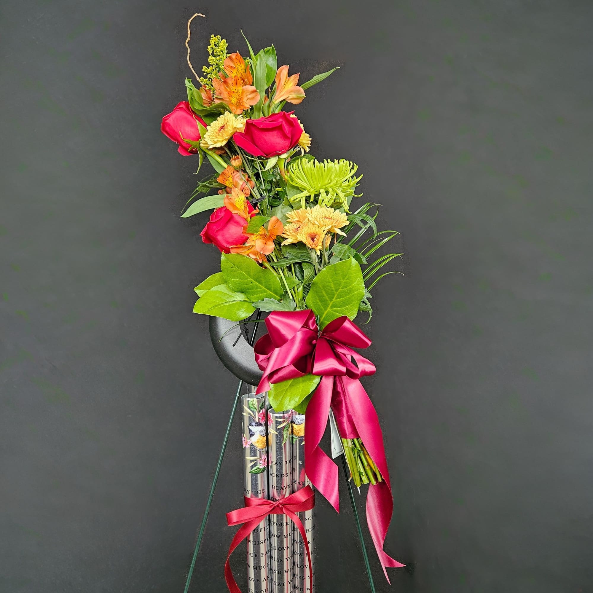 Tall floral spray with red roses, orange blooms, and a pink ribbon on a stand