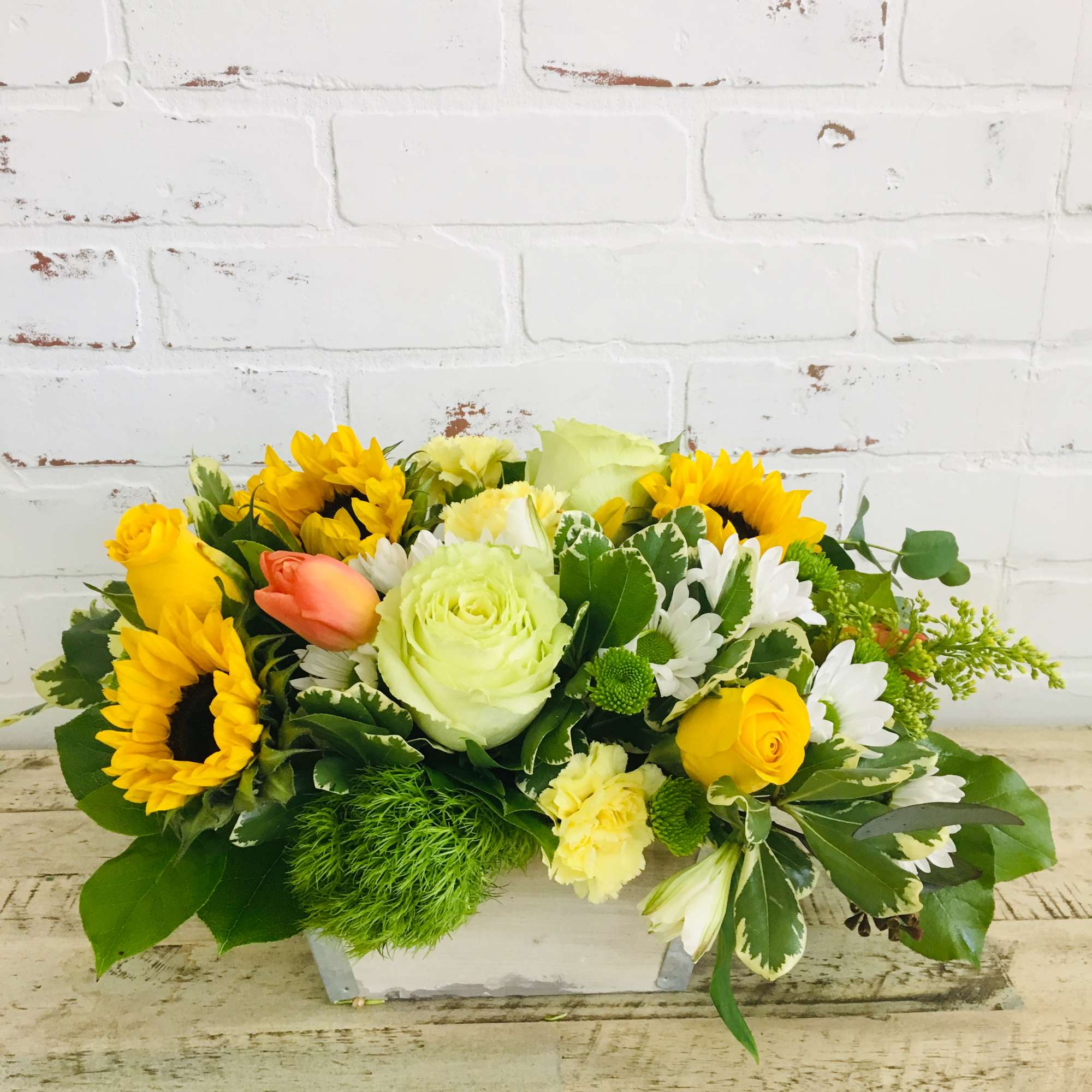 Low centerpiece of sunflowers, yellow roses, white daisies and green accents in a white wooden box.