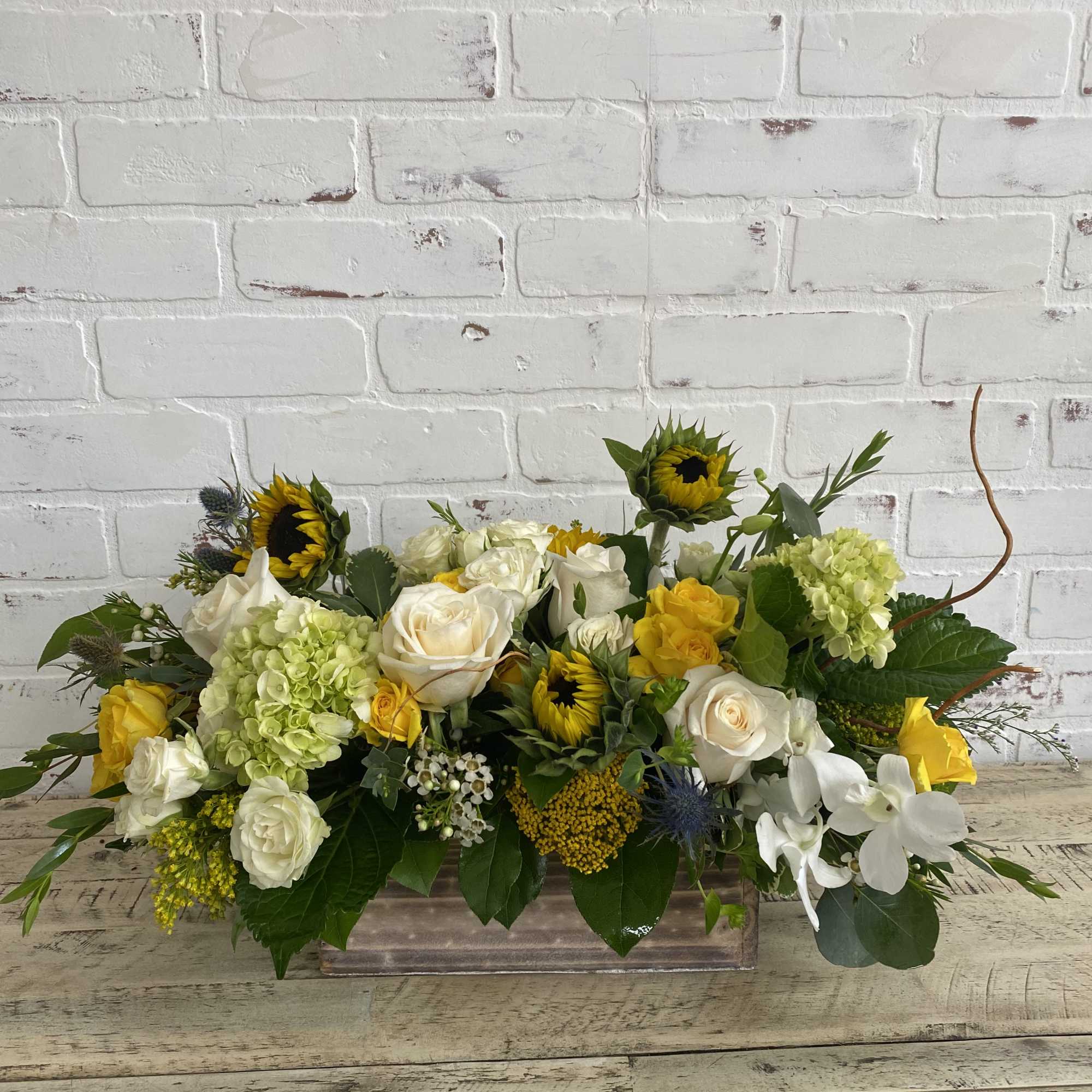 Low centerpiece of white and yellow roses, sunflowers, hydrangeas, and orchids in a rectangular container.