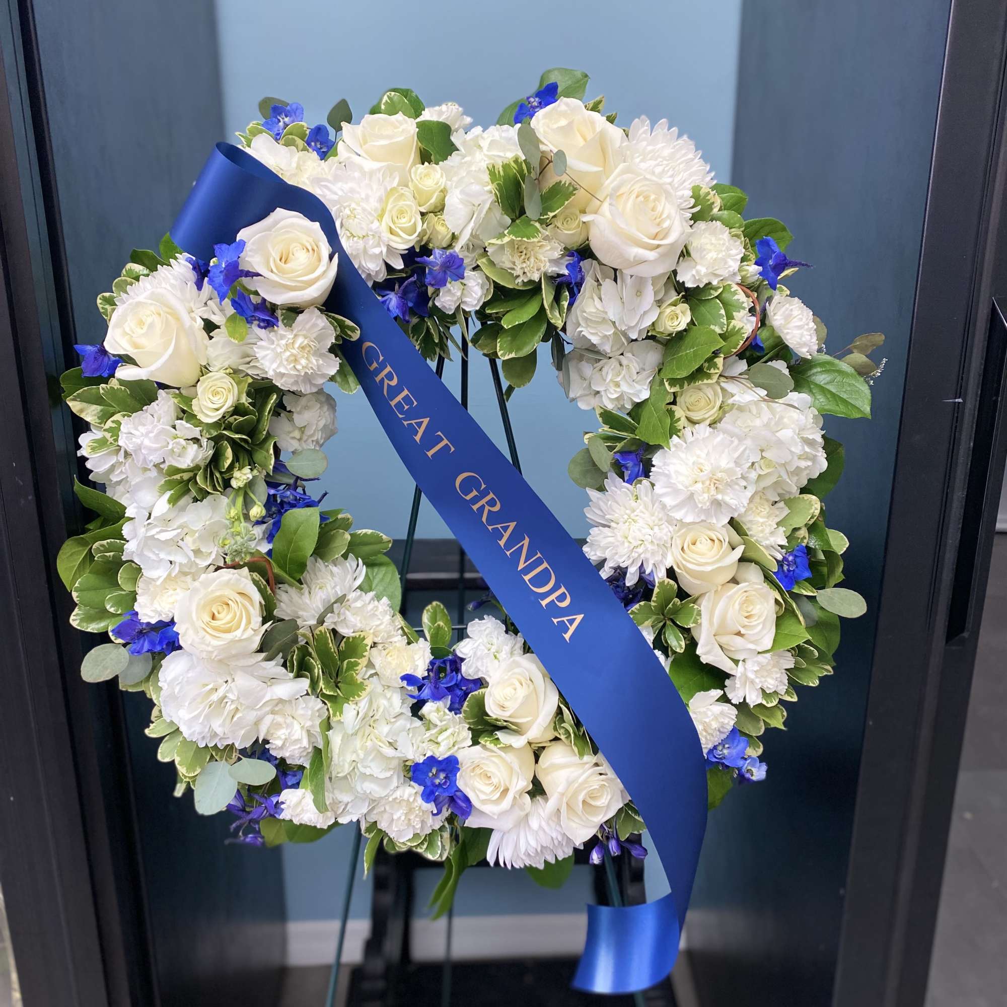 Heart-shaped white floral wreath with a blue ribbon on a stand