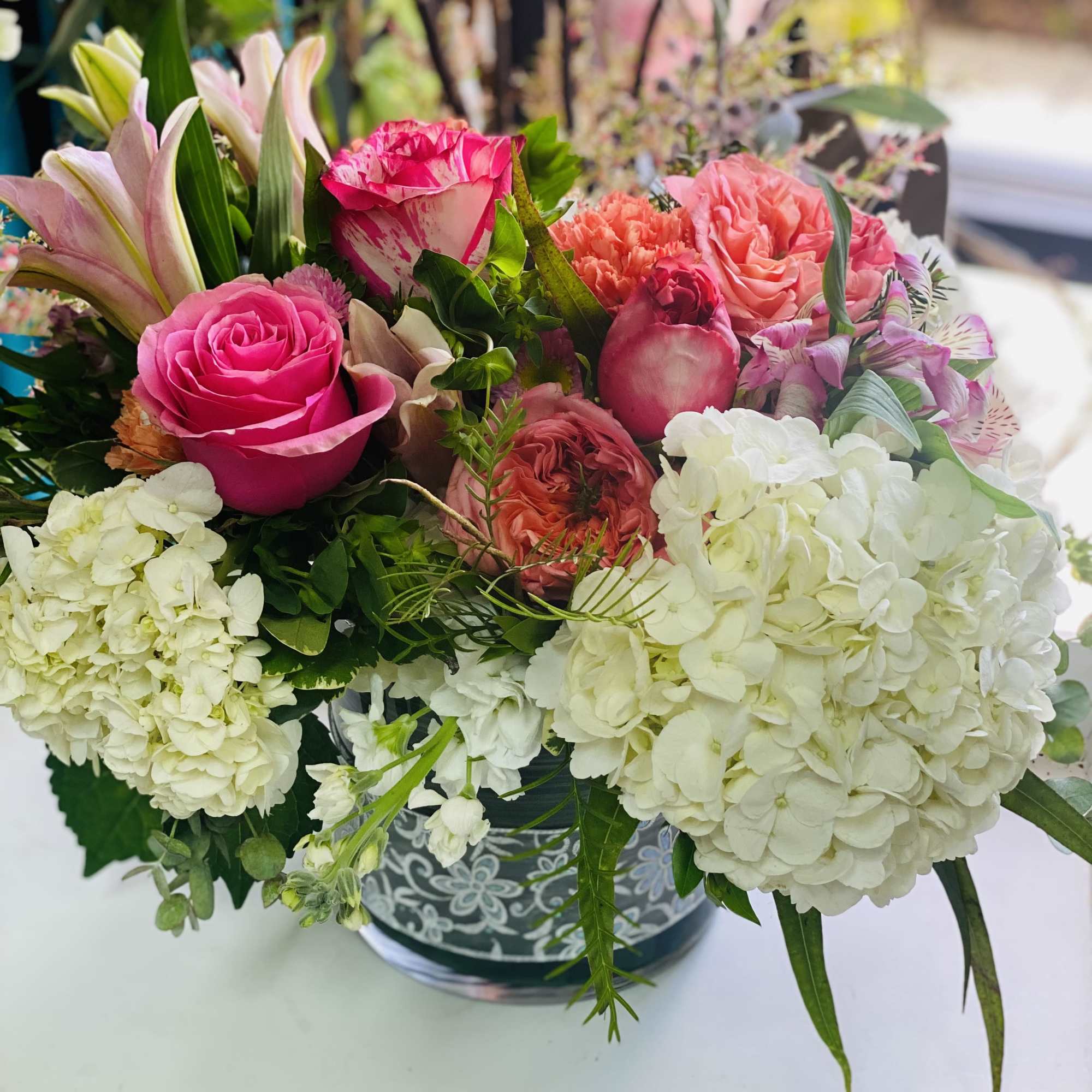 Low arrangement of white hydrangeas, pink roses, lilies, and mixed blooms in a patterned glass vase
