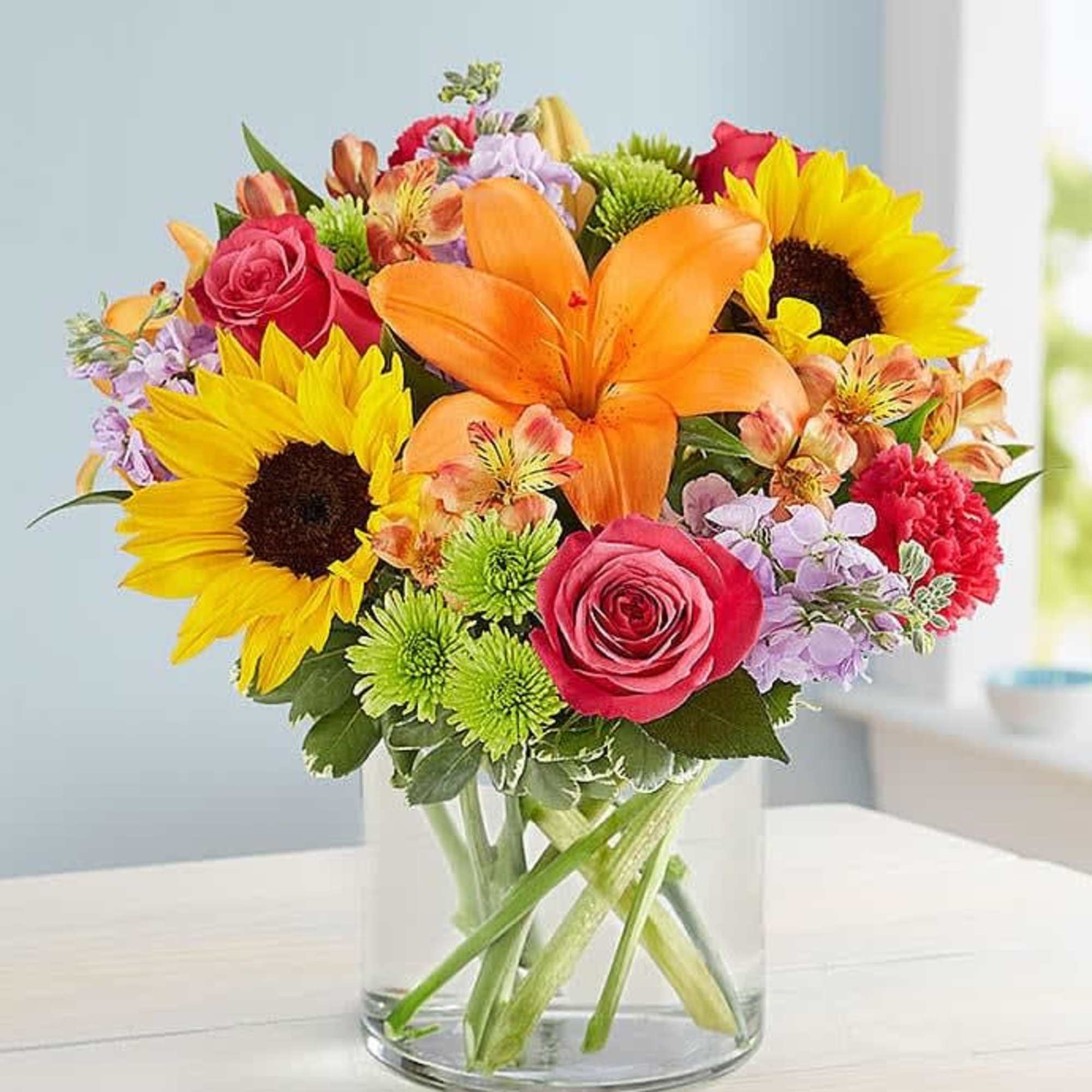 Bright mixed bouquet with sunflowers, orange lily, pink roses, and green mums in a clear glass vase