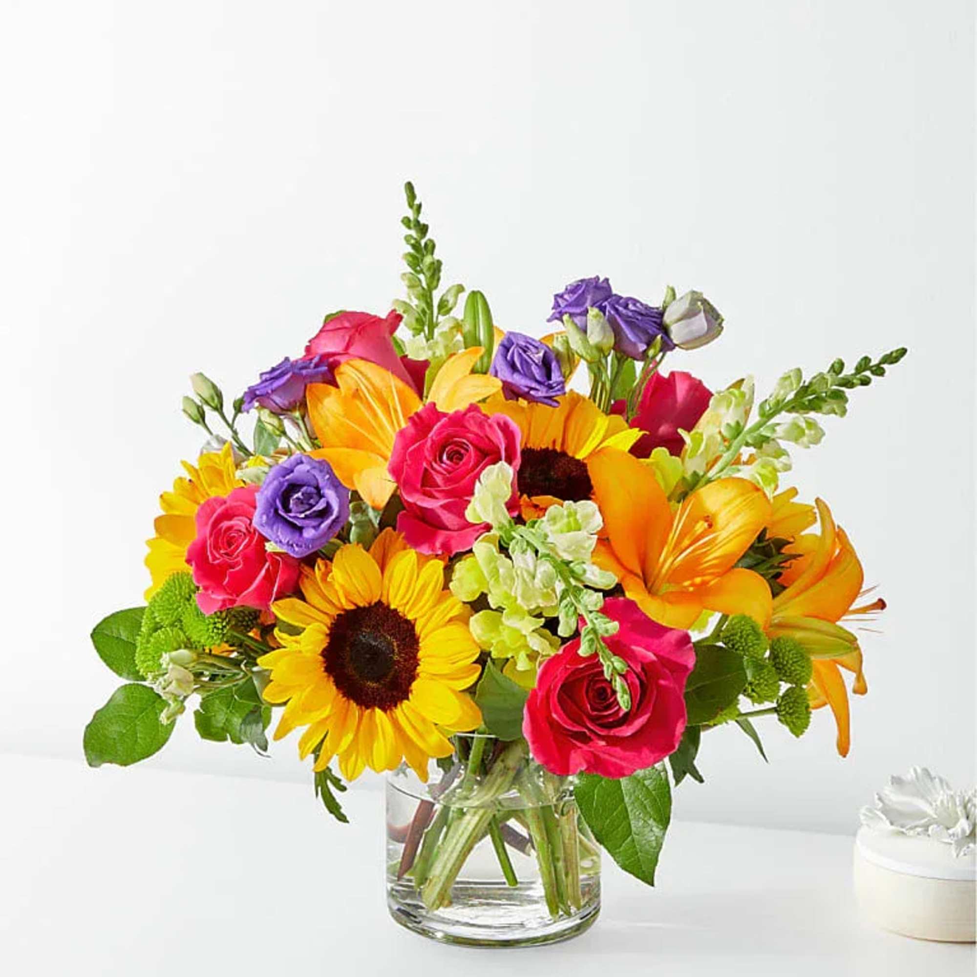 Bright mixed bouquet in a clear glass vase with sunflowers, roses, lilies, and purple blooms.
