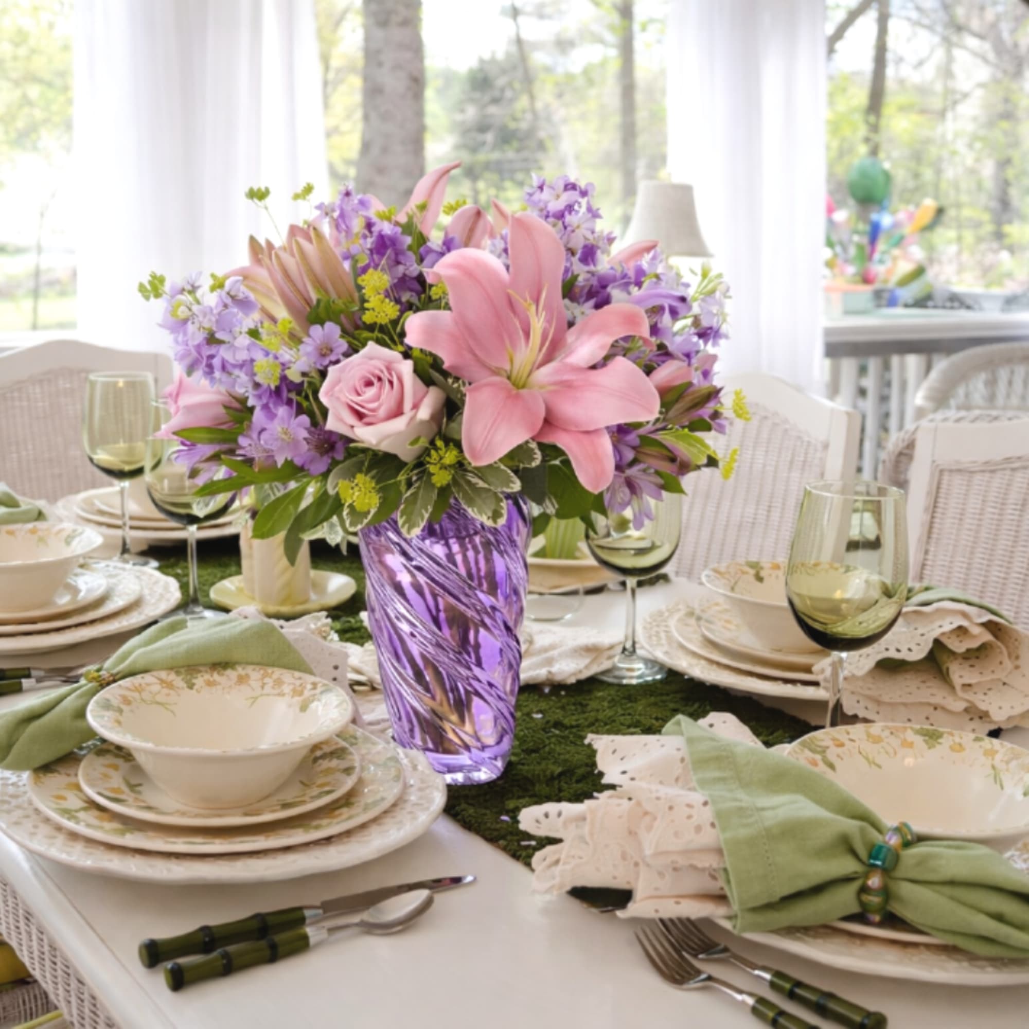Pink lilies and roses with lavender flowers in a purple glass vase on a set dining table