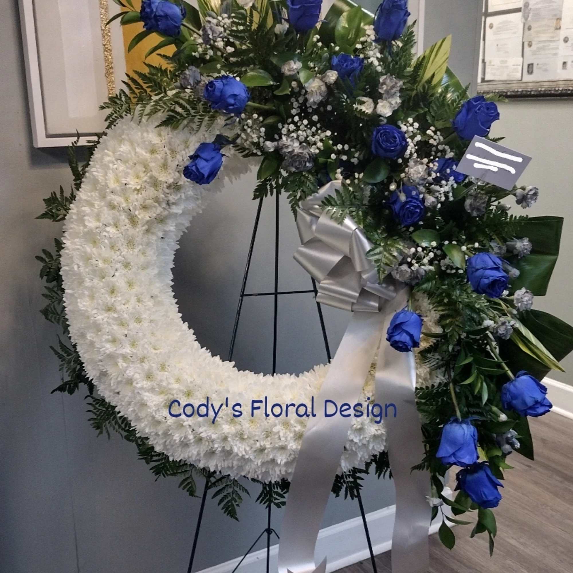 White floral wreath with blue roses on a stand and silver ribbon