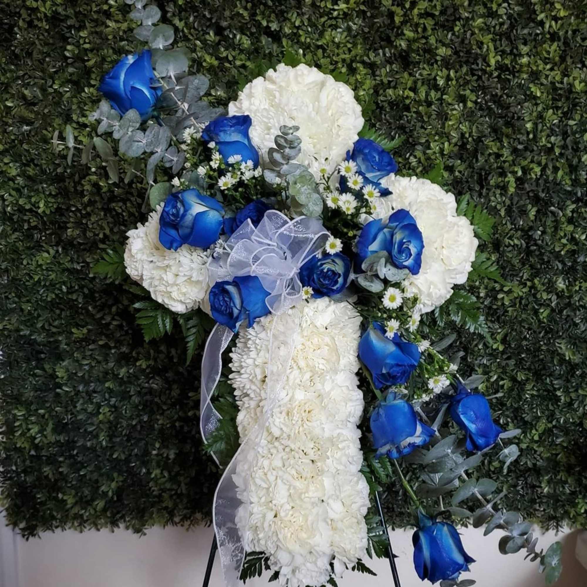 White floral cross with blue roses and ribbon on a greenery wall