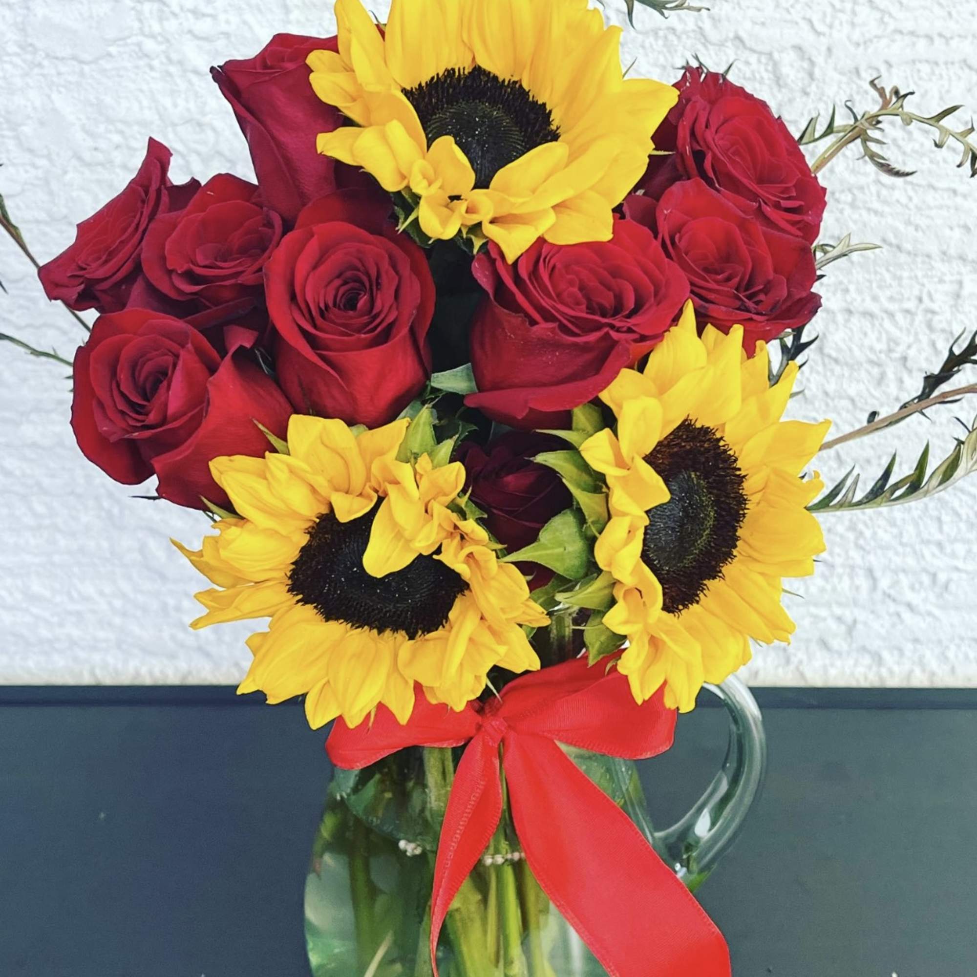 Red roses and yellow sunflowers in a clear glass pitcher with a red ribbon
