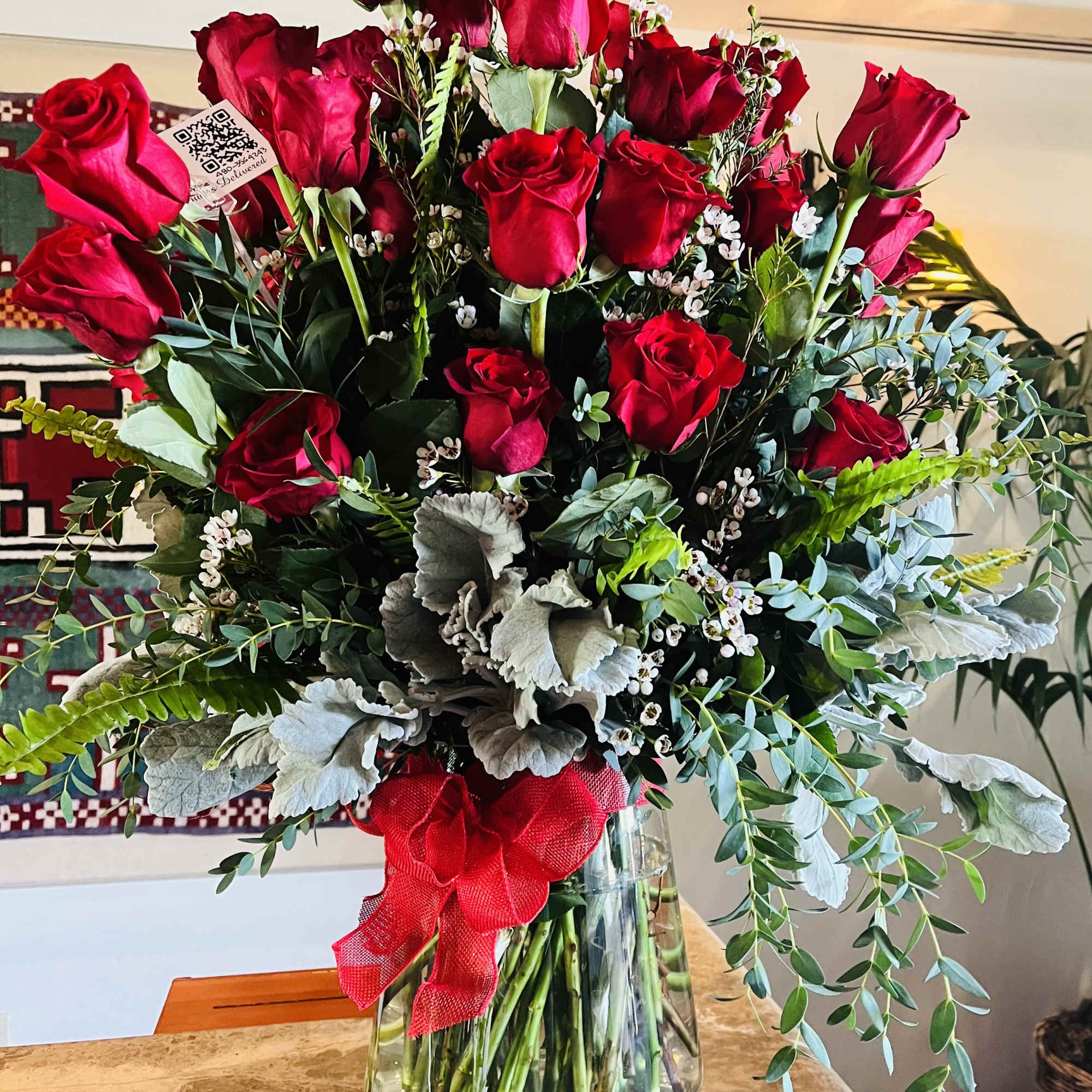 Red roses arranged in a clear glass vase with greenery and a red ribbon