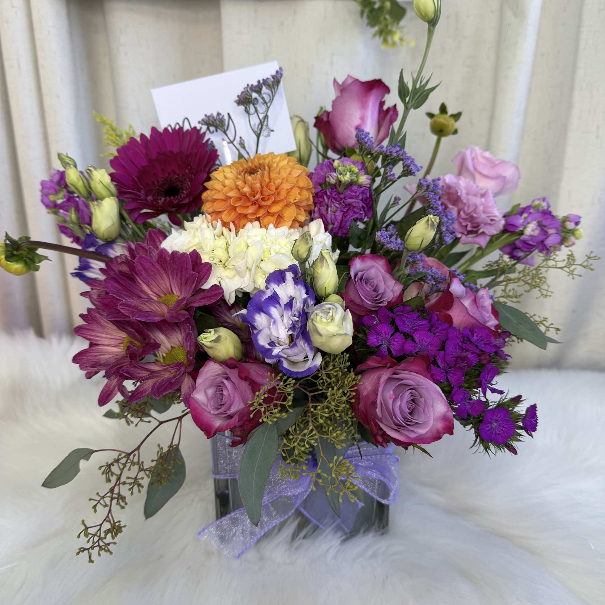 Mixed bouquet of purple, pink, orange, and white flowers in a square vase with ribbon.