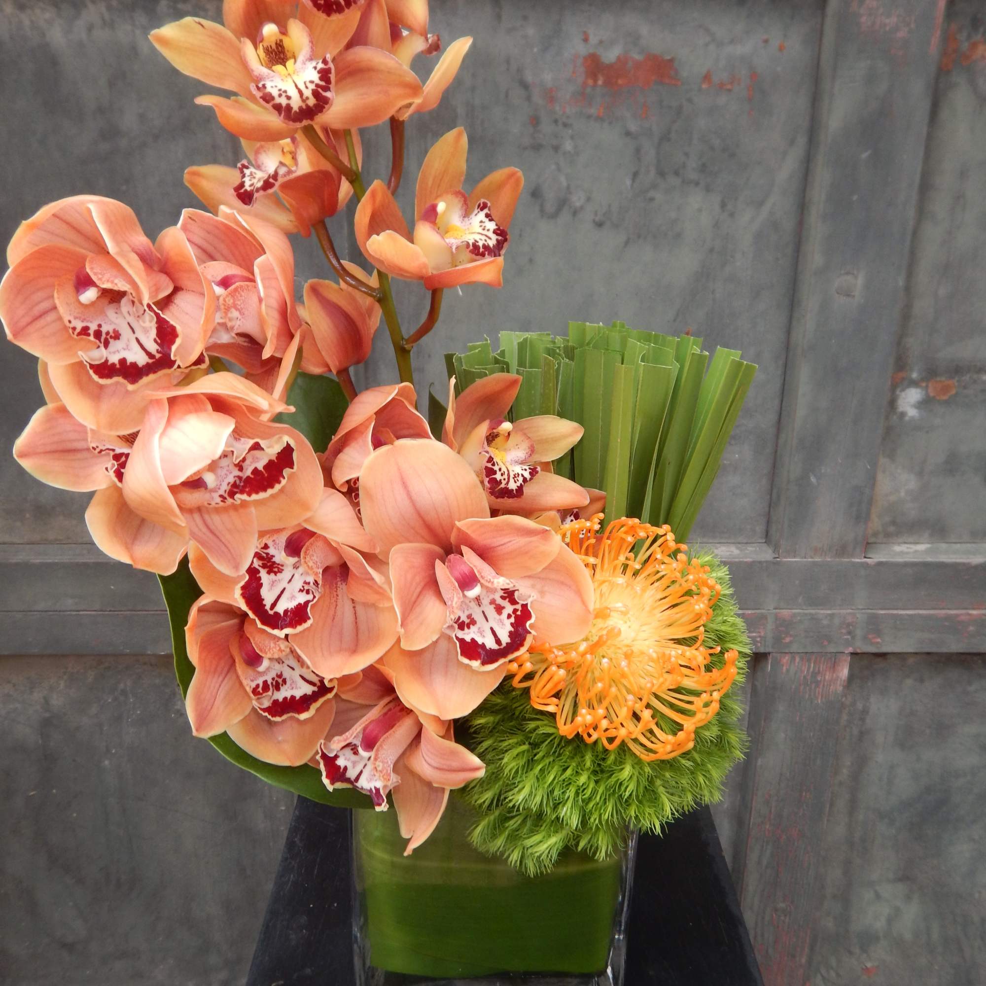 Orange orchids arranged in a square glass vase with green accents