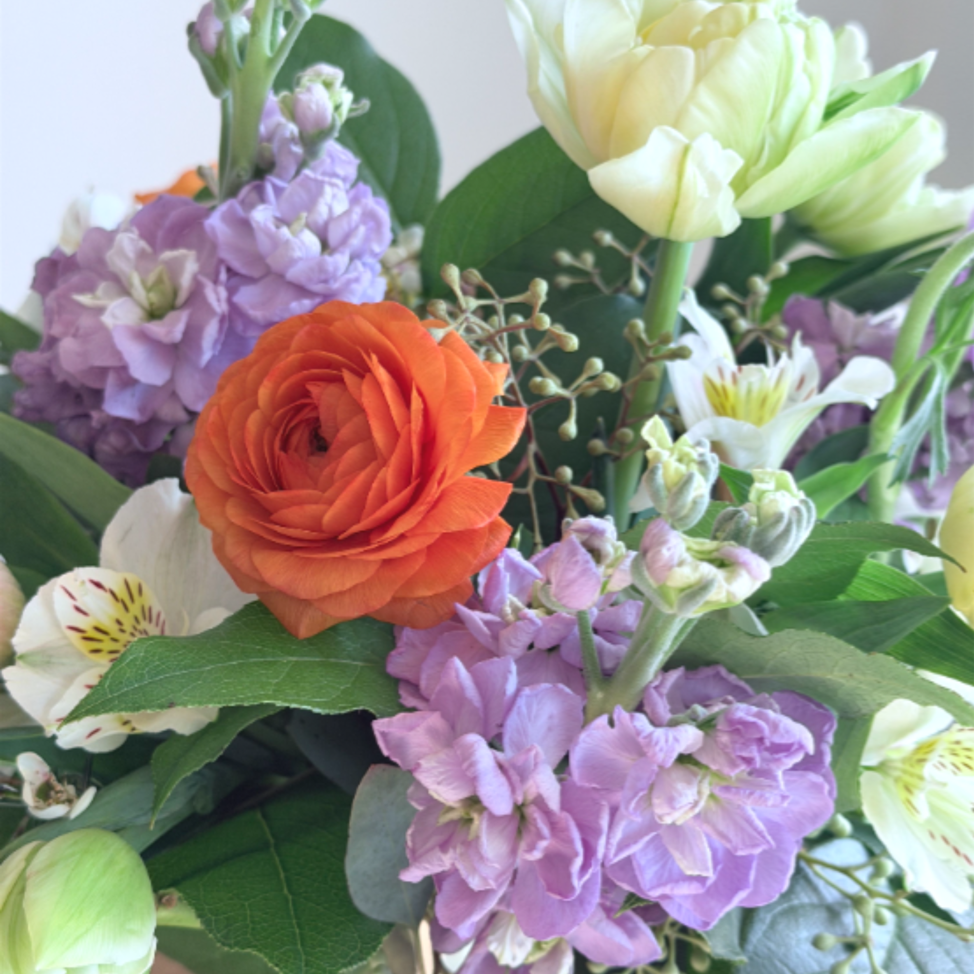 Mixed bouquet of orange ranunculus, pale tulips, and lavender blooms in a clear glass vase