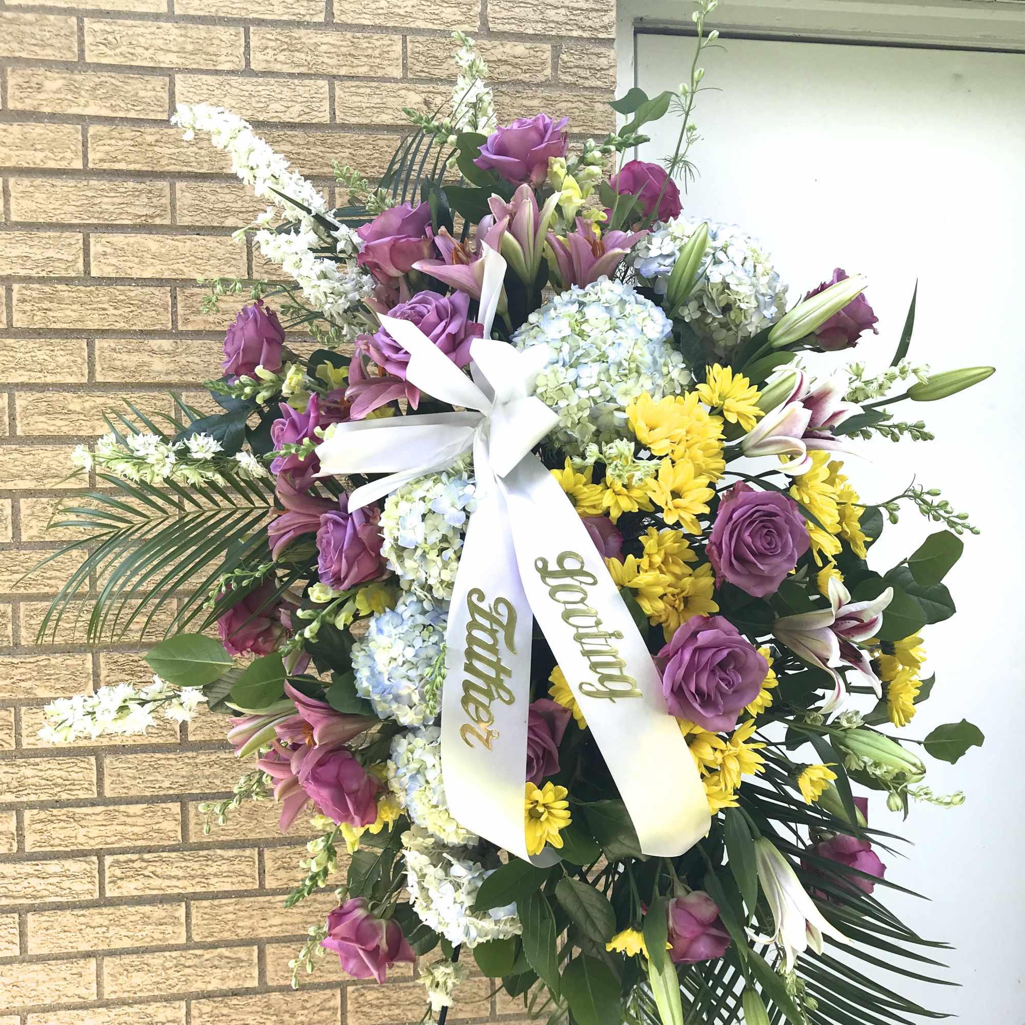 Standing sympathy spray with purple roses, lilies, hydrangeas, and yellow daisies on a white ribbon easel