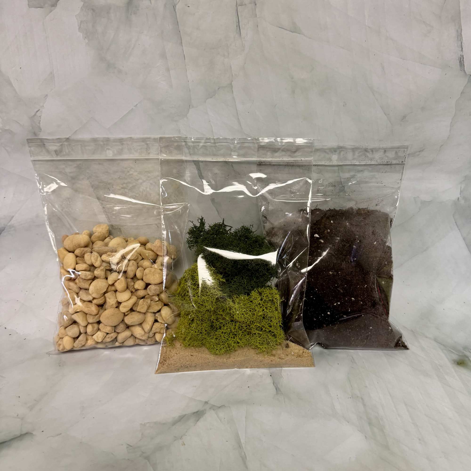 Three clear bags of decorative stones, moss, and dark soil on a light surface