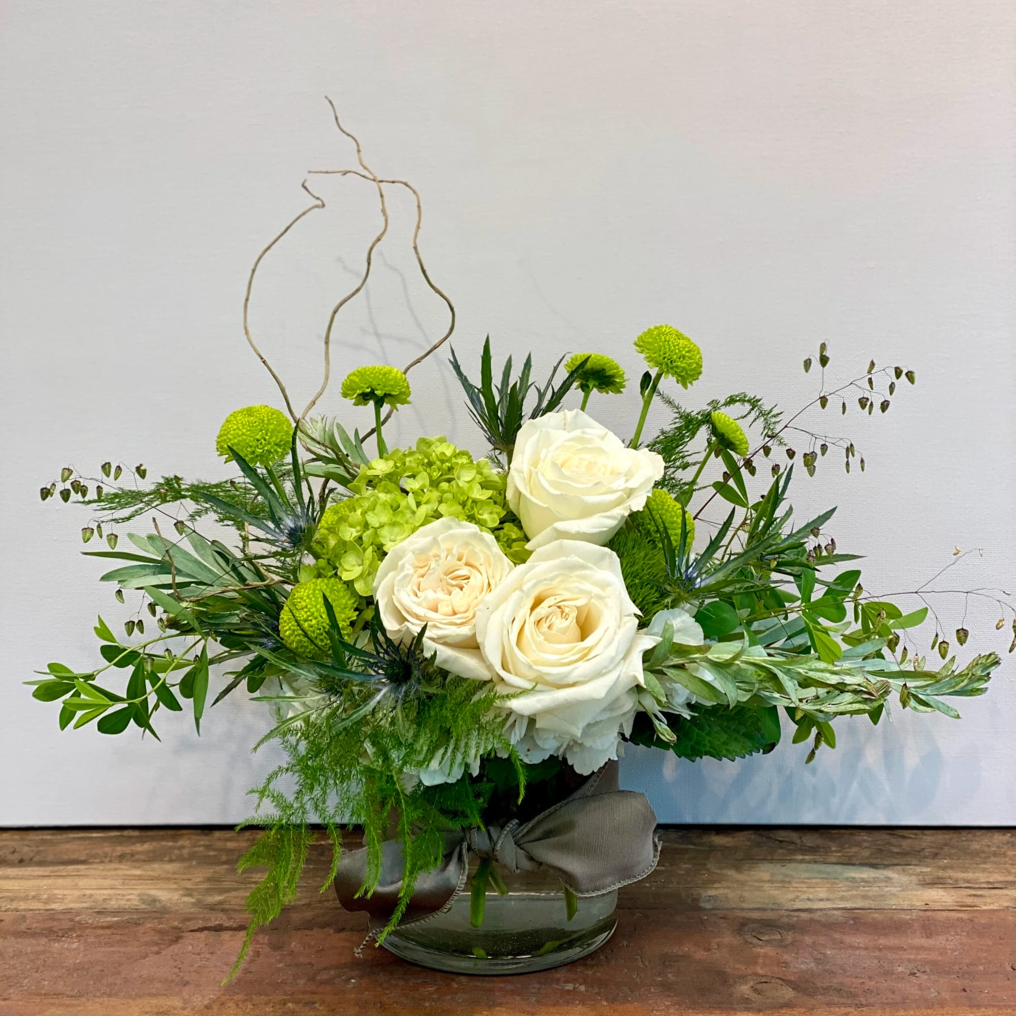 Low glass vase of white roses, green hydrangea, and green button poms with airy branches and foliage.