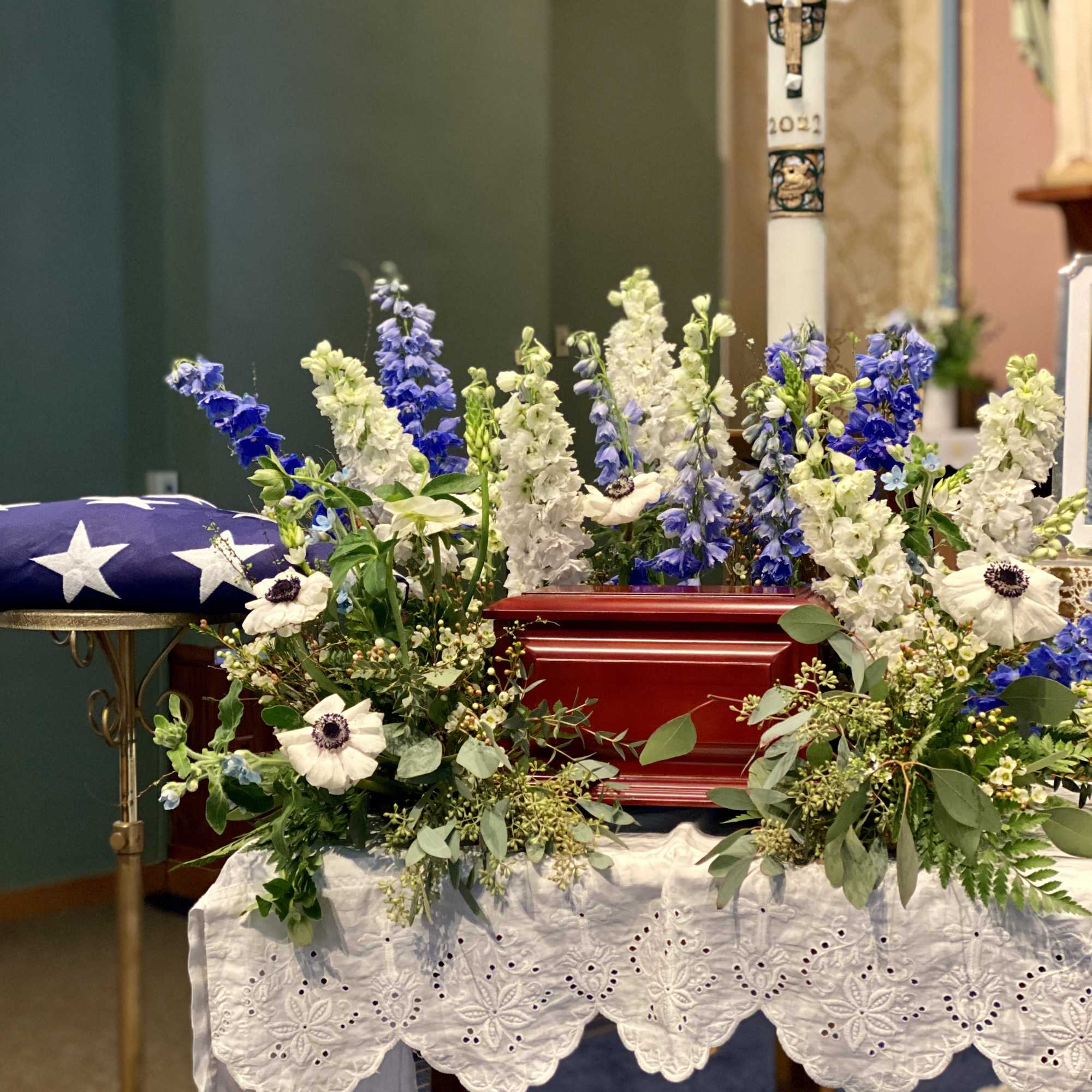 Patriotic funeral casket spray of blue and white flowers beside a folded American flag.
