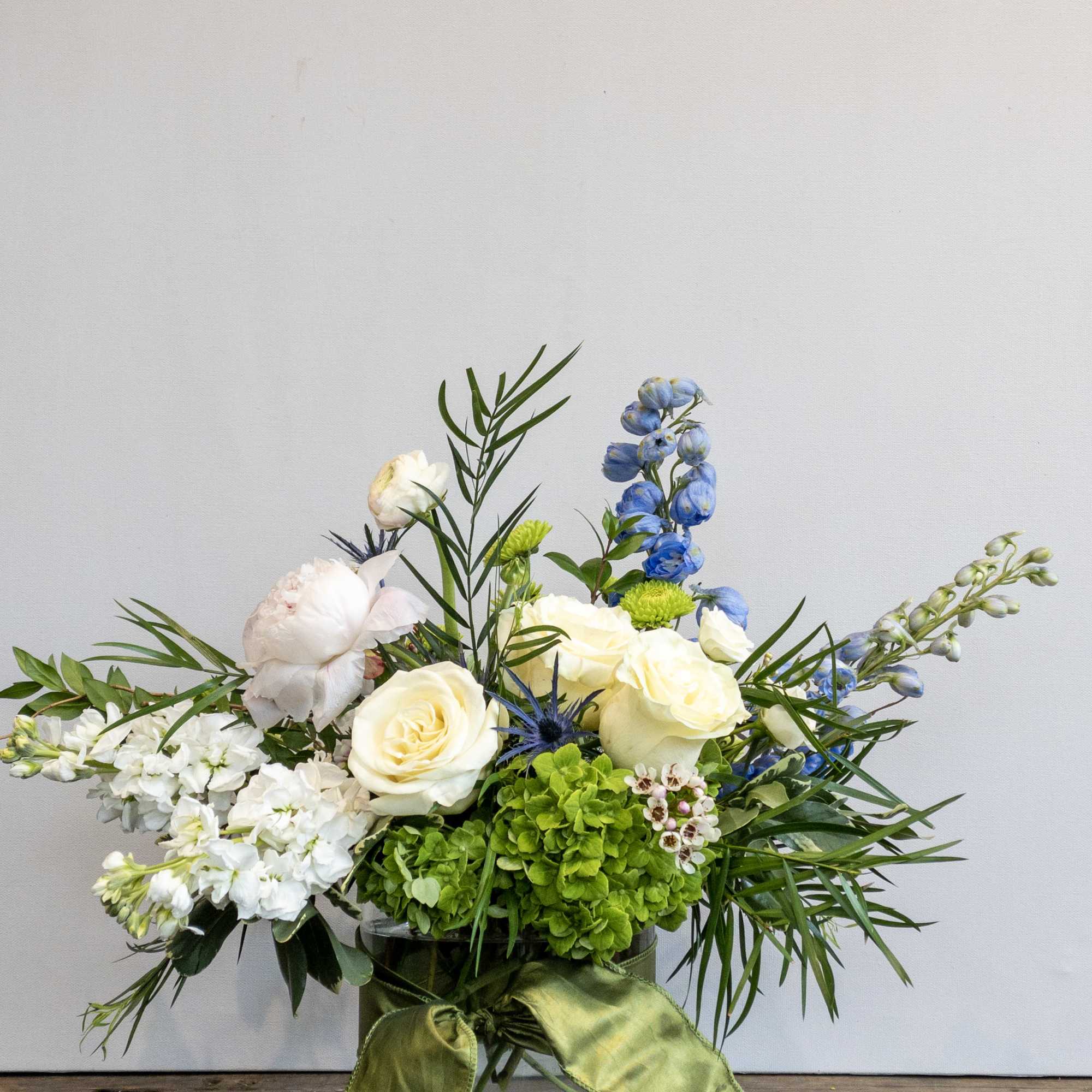 Low arrangement of white roses, green hydrangea, and blue delphinium in a glass vase with ribbon