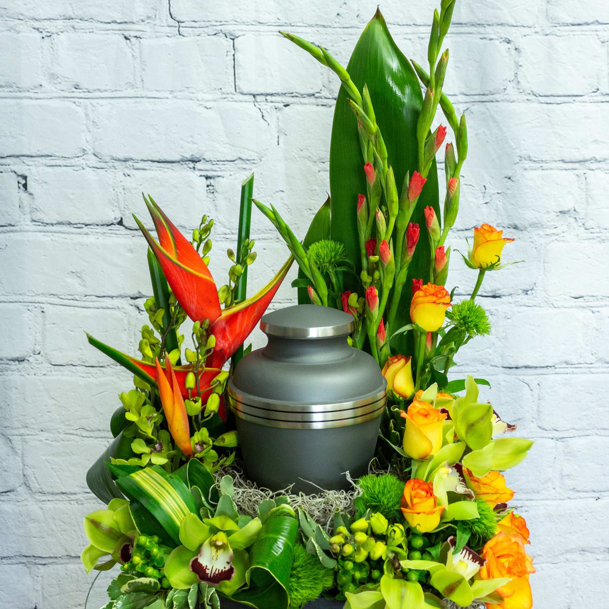 Cremation urn surrounded by orange roses, green orchids, and tropical flowers in a tall arrangement