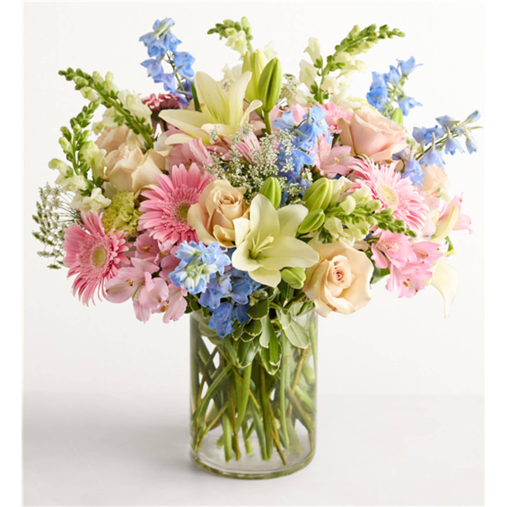 Mixed pastel bouquet in a clear glass vase with lilies, roses, gerberas, and blue flowers