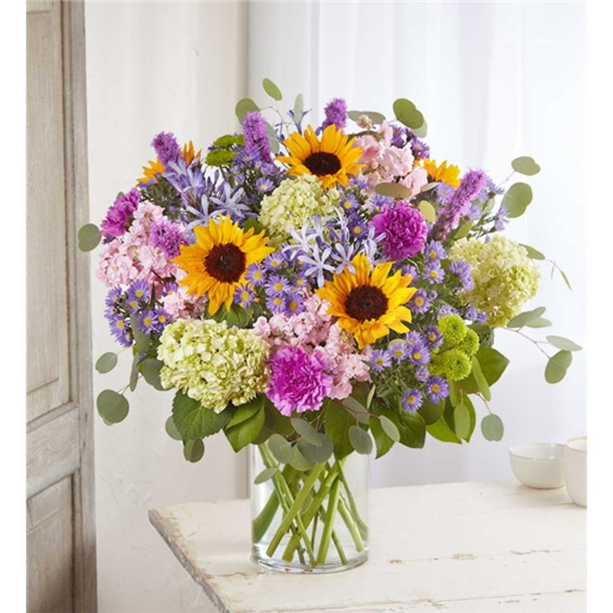 Mixed bouquet with sunflowers, purple blooms, and pink flowers in a clear glass vase