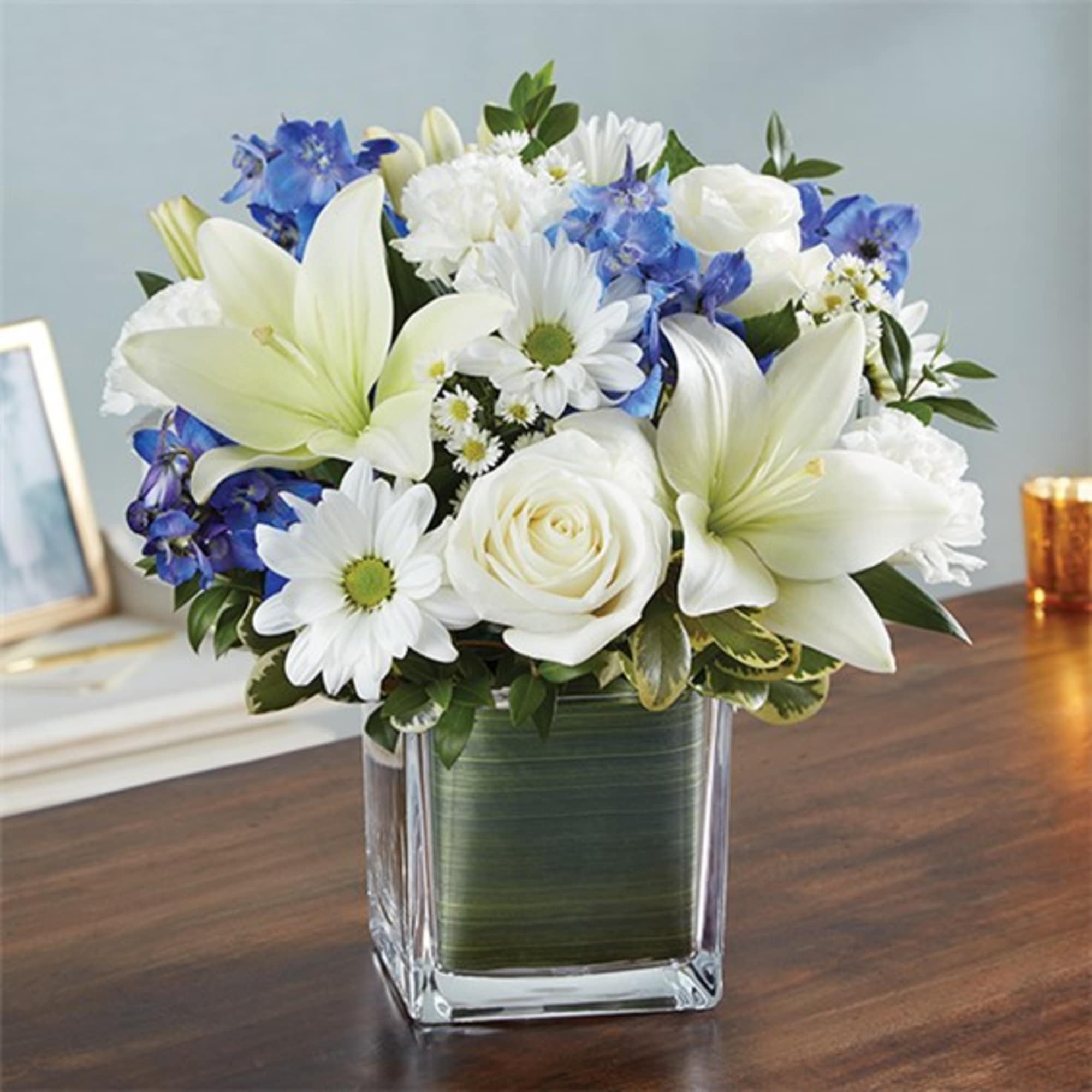White lilies, roses, and blue flowers arranged in a square glass vase