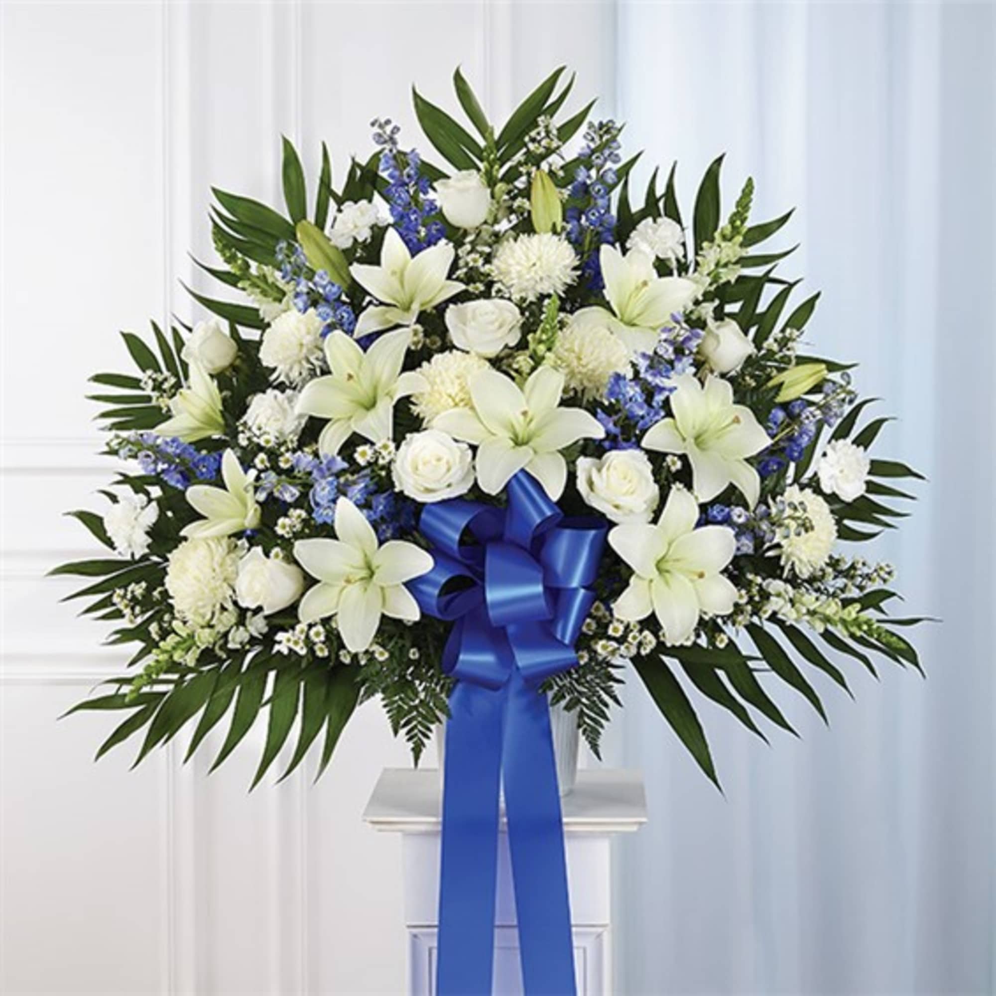 White lilies and roses arranged with blue flowers in a vase, tied with a blue ribbon.