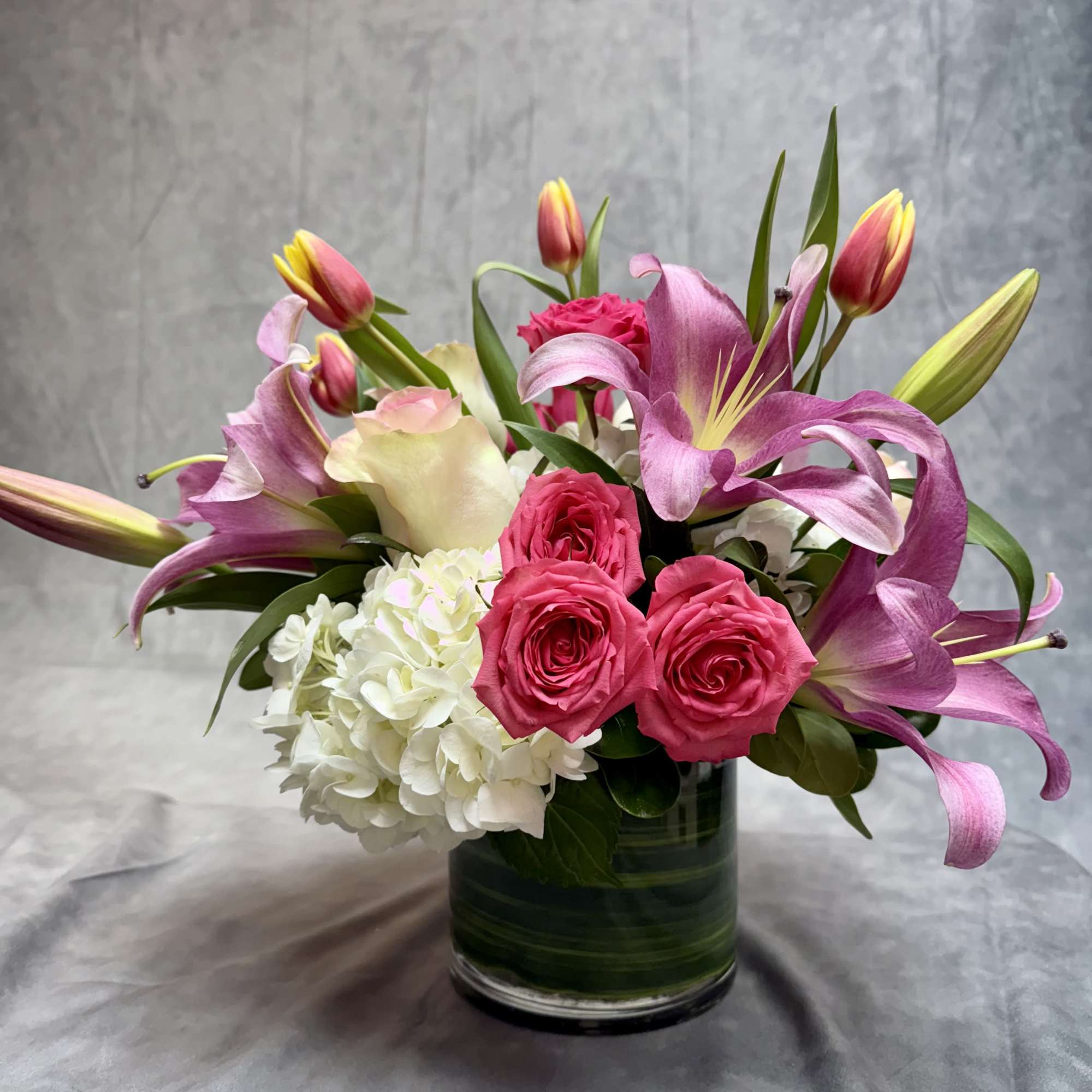 Pink lilies and roses arranged in a glass vase with white hydrangea.
