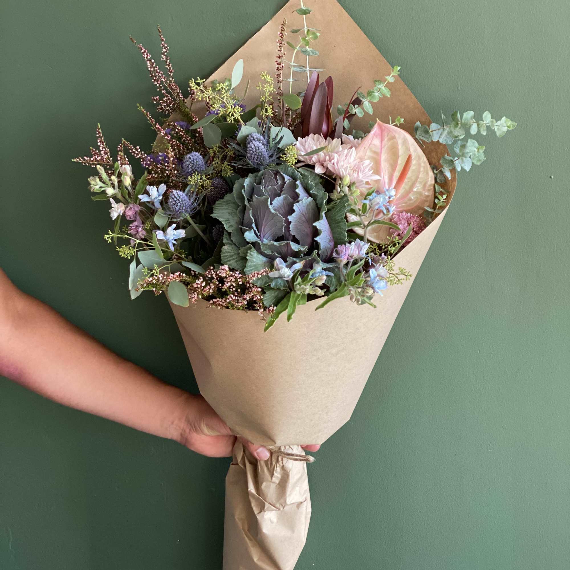 Handheld bouquet with anthurium, thistle, and mixed blooms in kraft paper