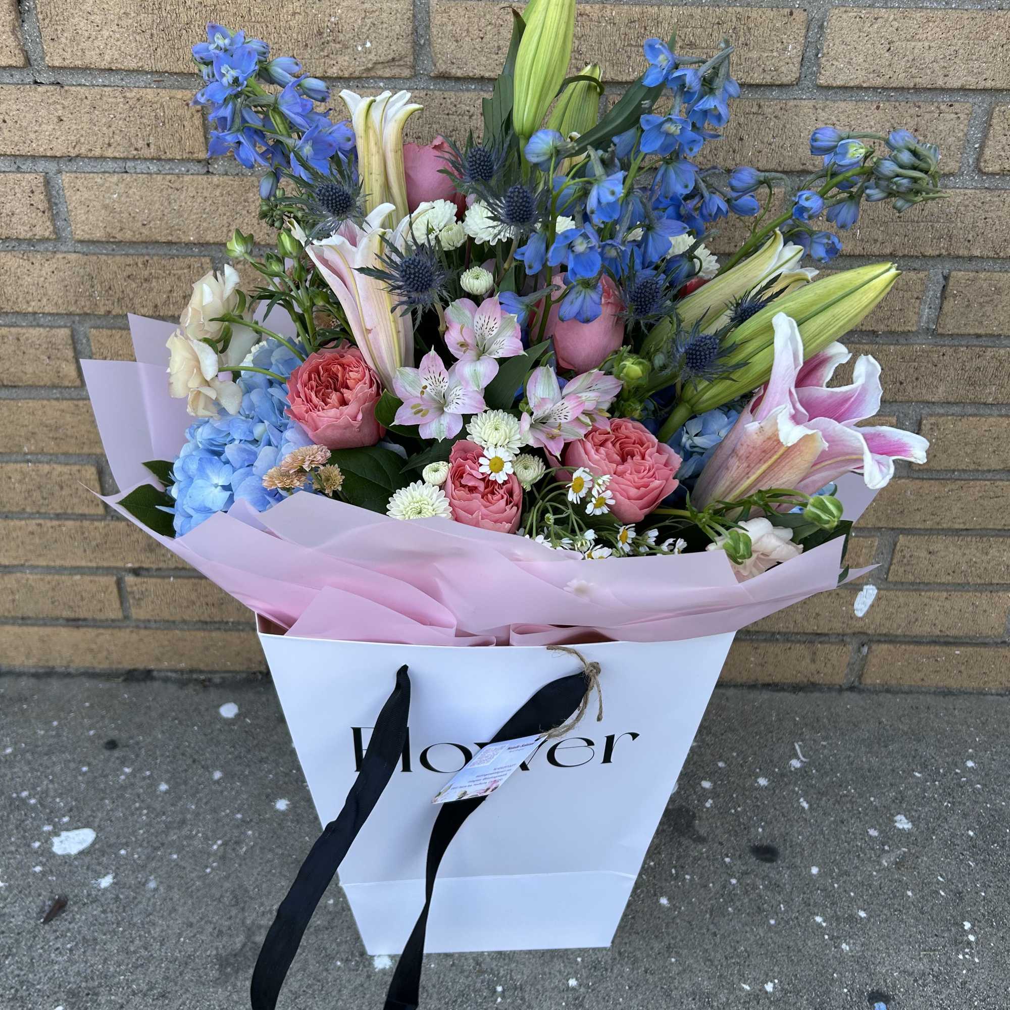 Mixed bouquet with blue delphinium, pink roses, lilies, and hydrangea in a gift bag