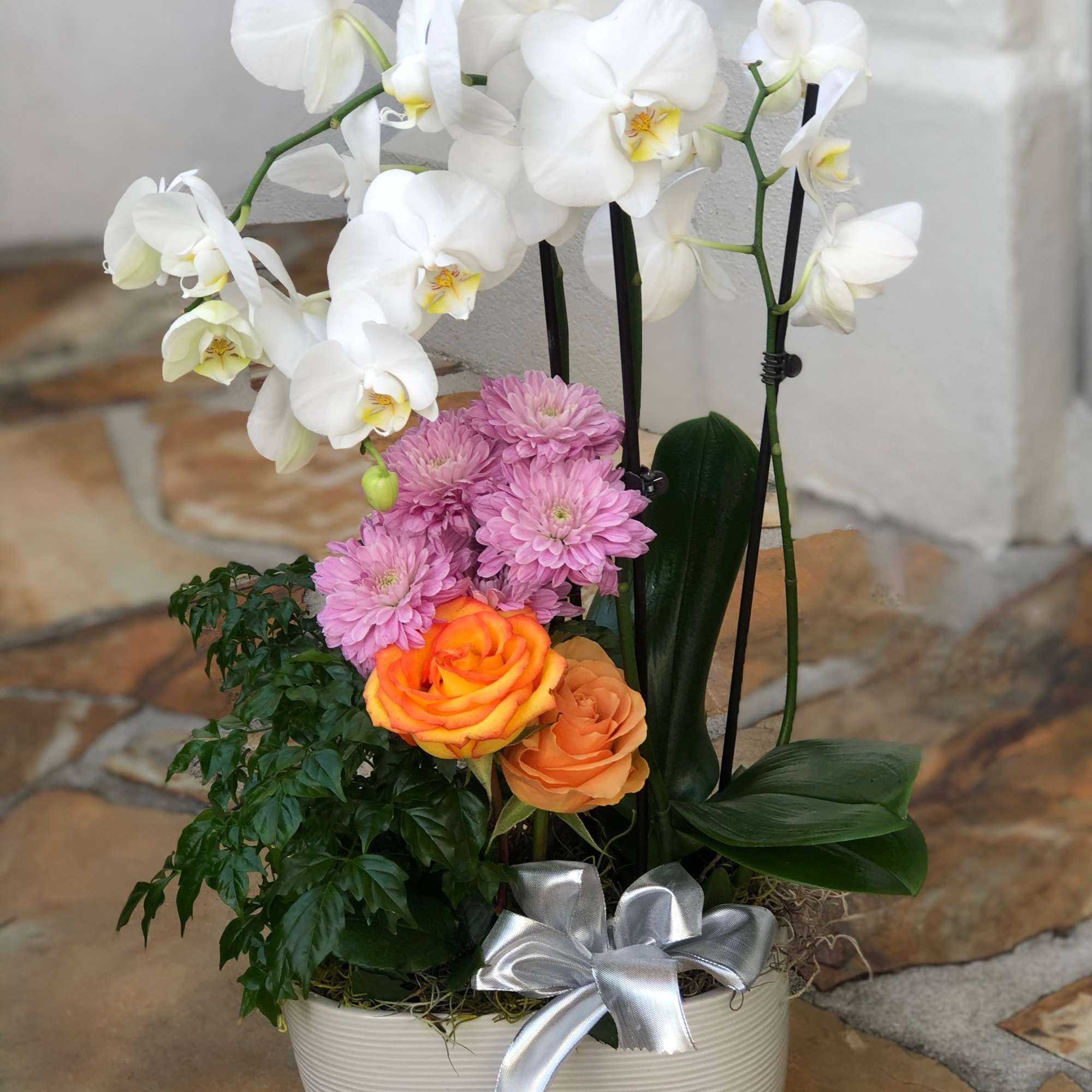 White orchid plant with pink mums and orange roses in a white ceramic planter with a silver bow