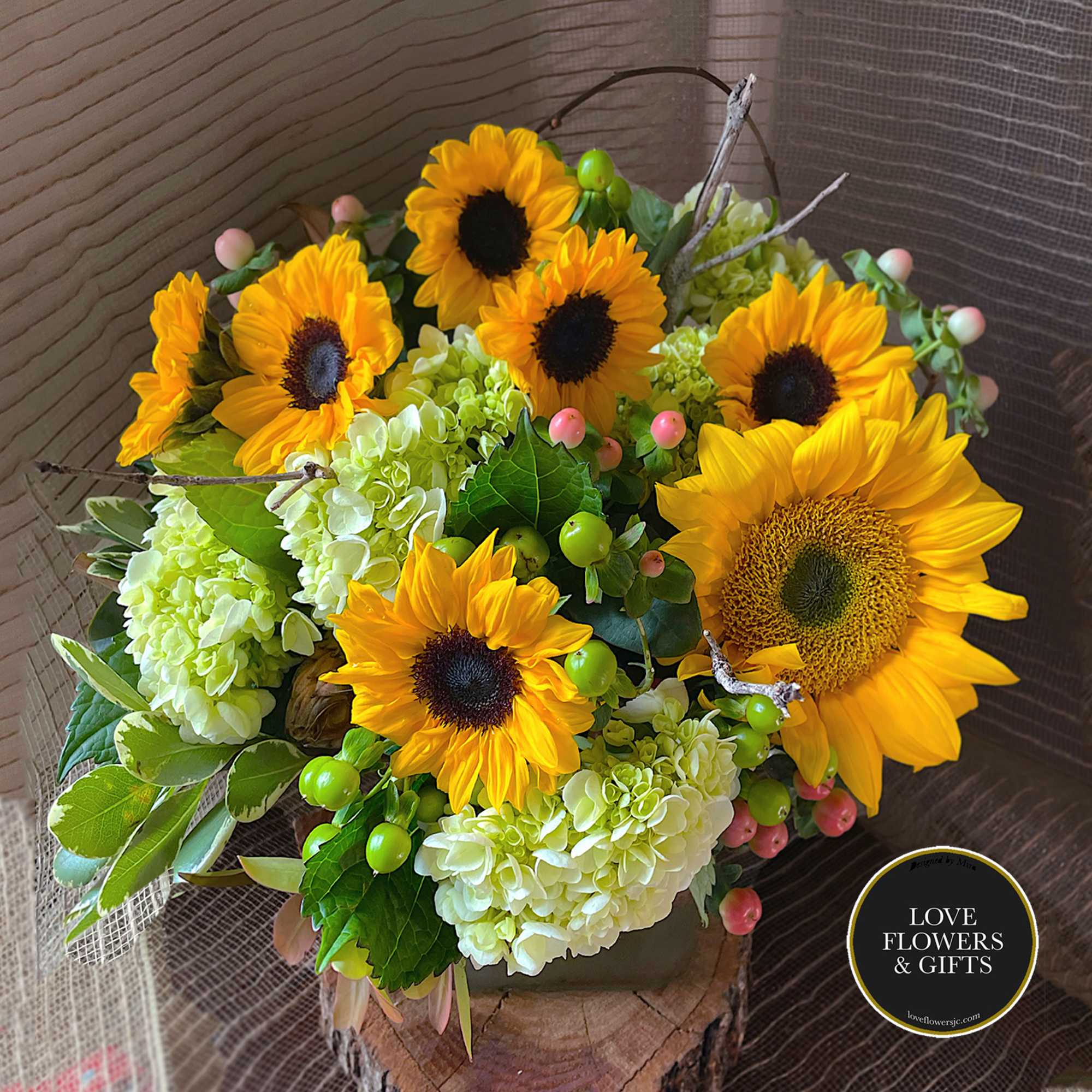 Arrangement of yellow sunflowers and pale green hydrangeas in a low vase on a wood slice