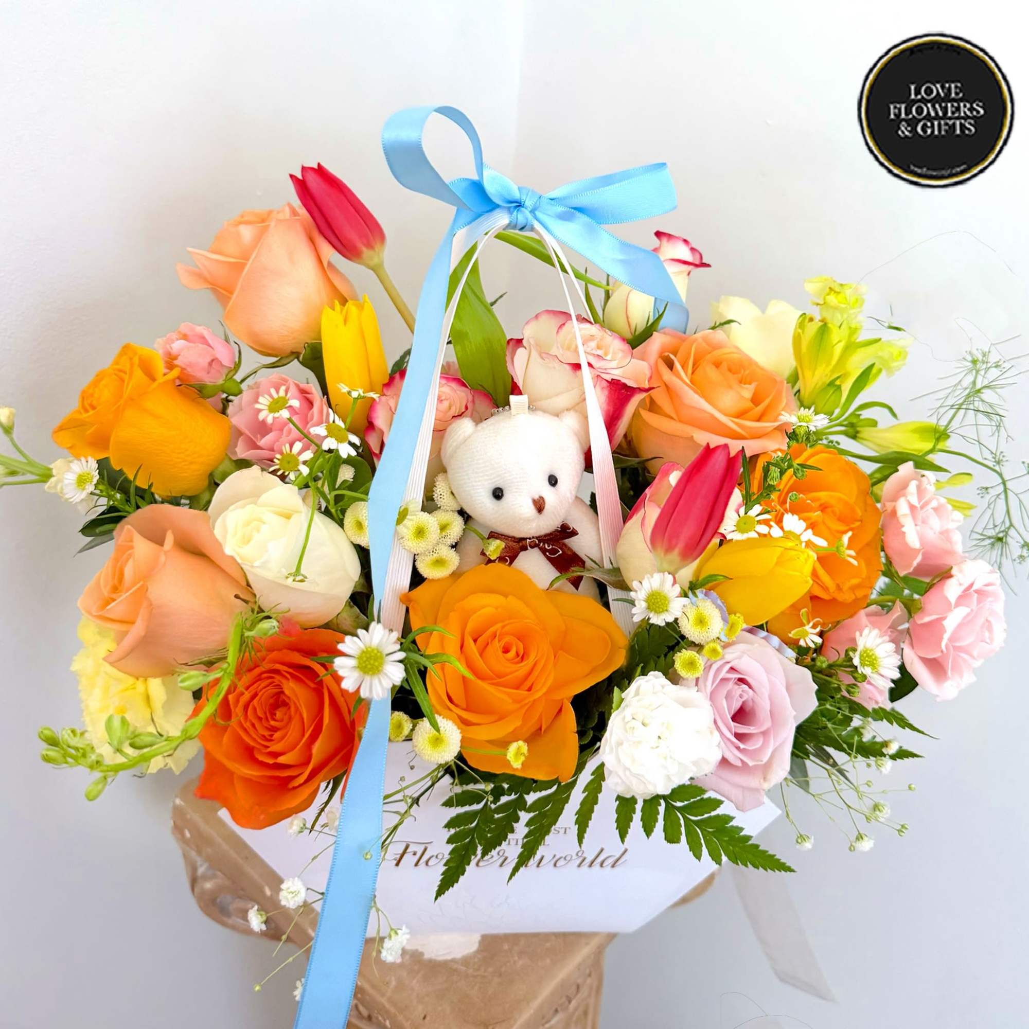 Bright mixed arrangement of roses and tulips with a small white teddy bear in a white box.