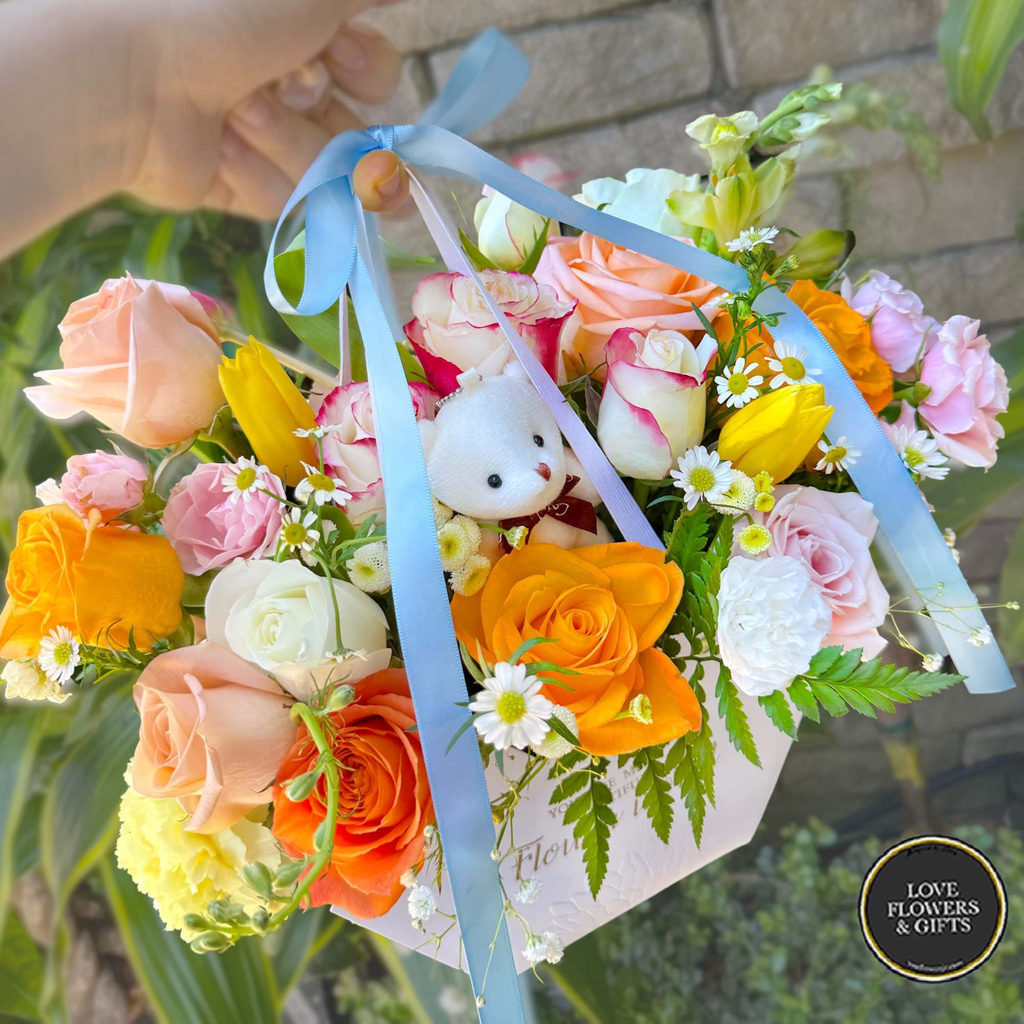 Colorful arrangement of roses and mixed flowers in a box with blue ribbons and a small white plush bear.