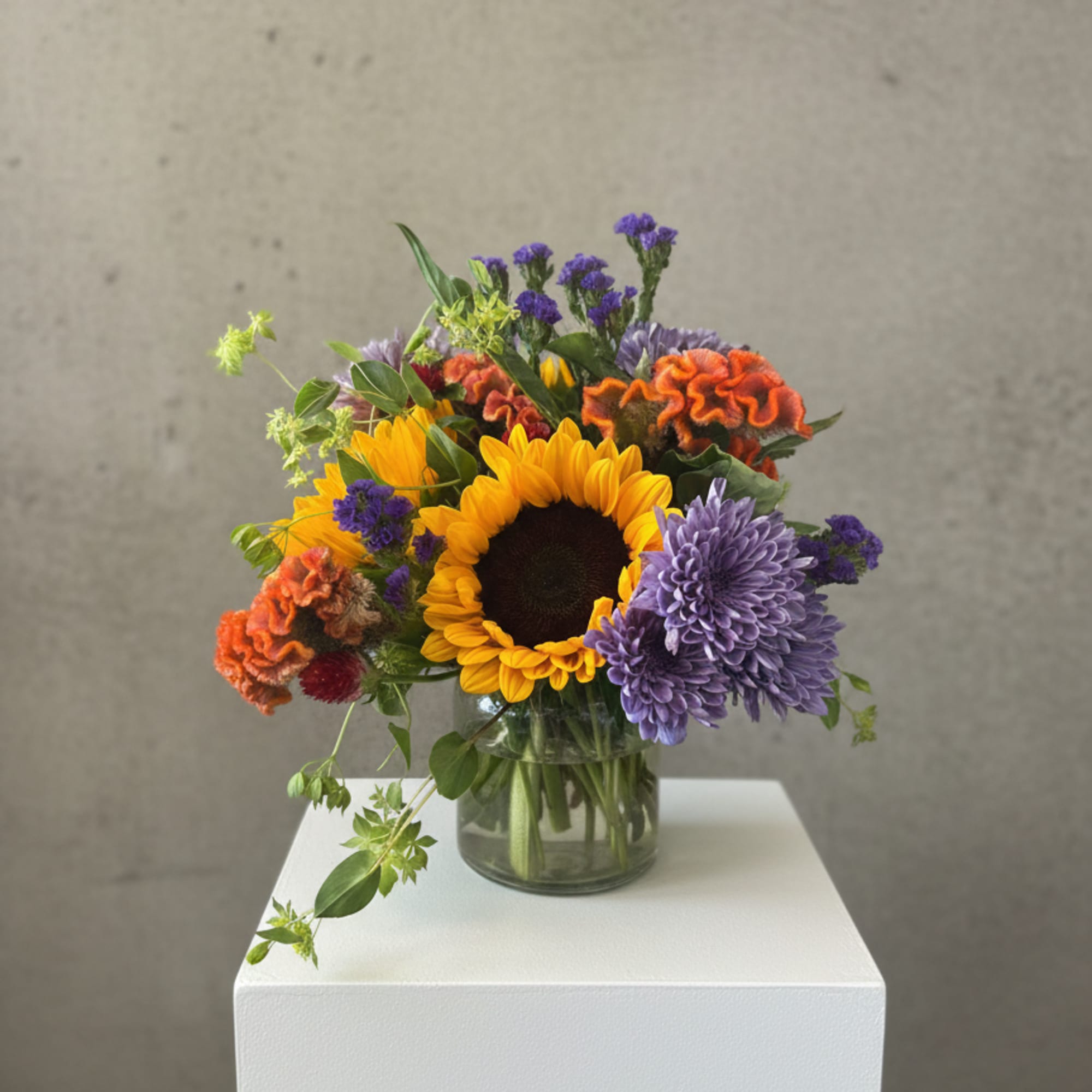 Bouquet of sunflowers, purple mums, and orange celosia in a glass vase