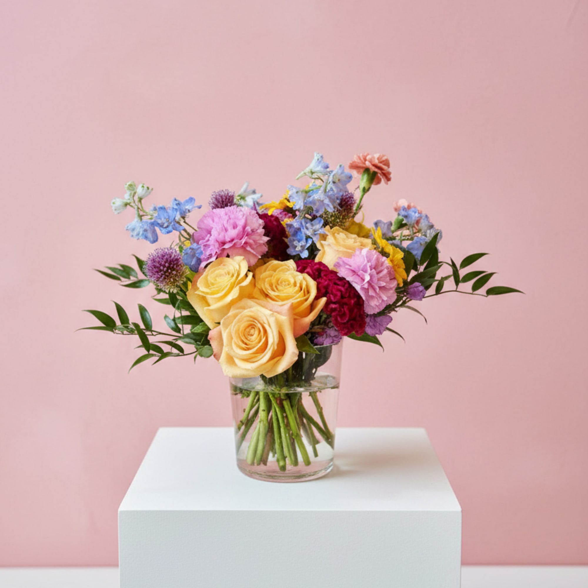 Mixed bouquet of yellow roses, pink blooms, and blue flowers in a clear glass vase