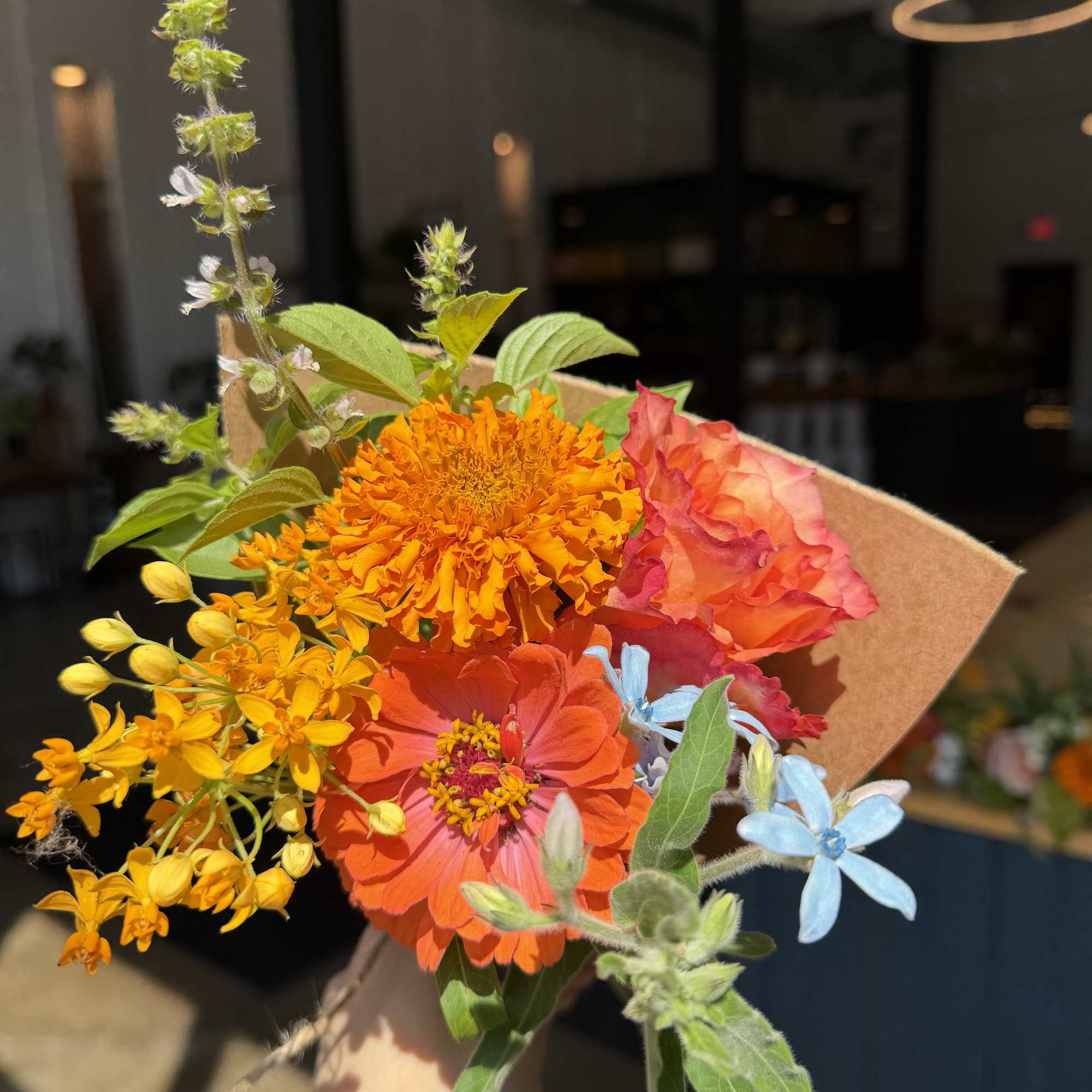 Handheld bouquet with orange, yellow, pink, and blue flowers wrapped in kraft paper.