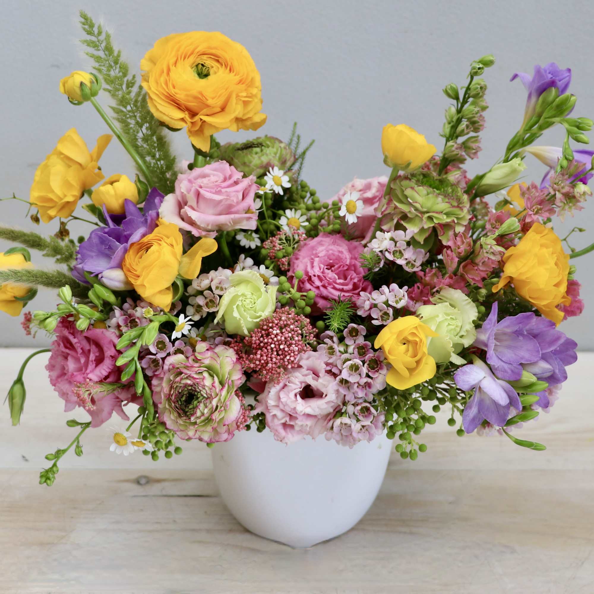 Low arrangement of yellow ranunculus, pink blooms, and purple flowers in a white bowl vase
