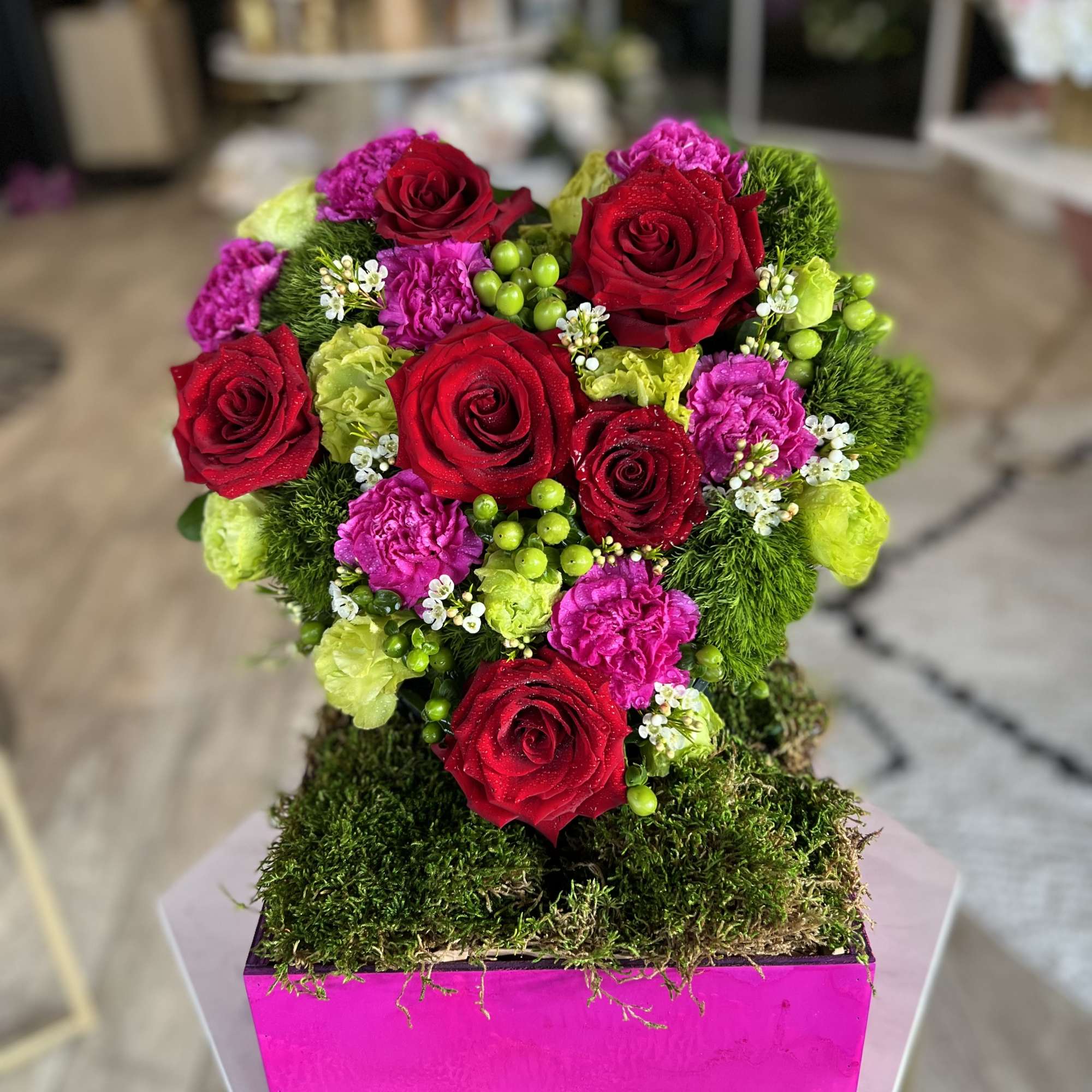 Heart-shaped arrangement of red roses, pink carnations, and green accents in a pink box