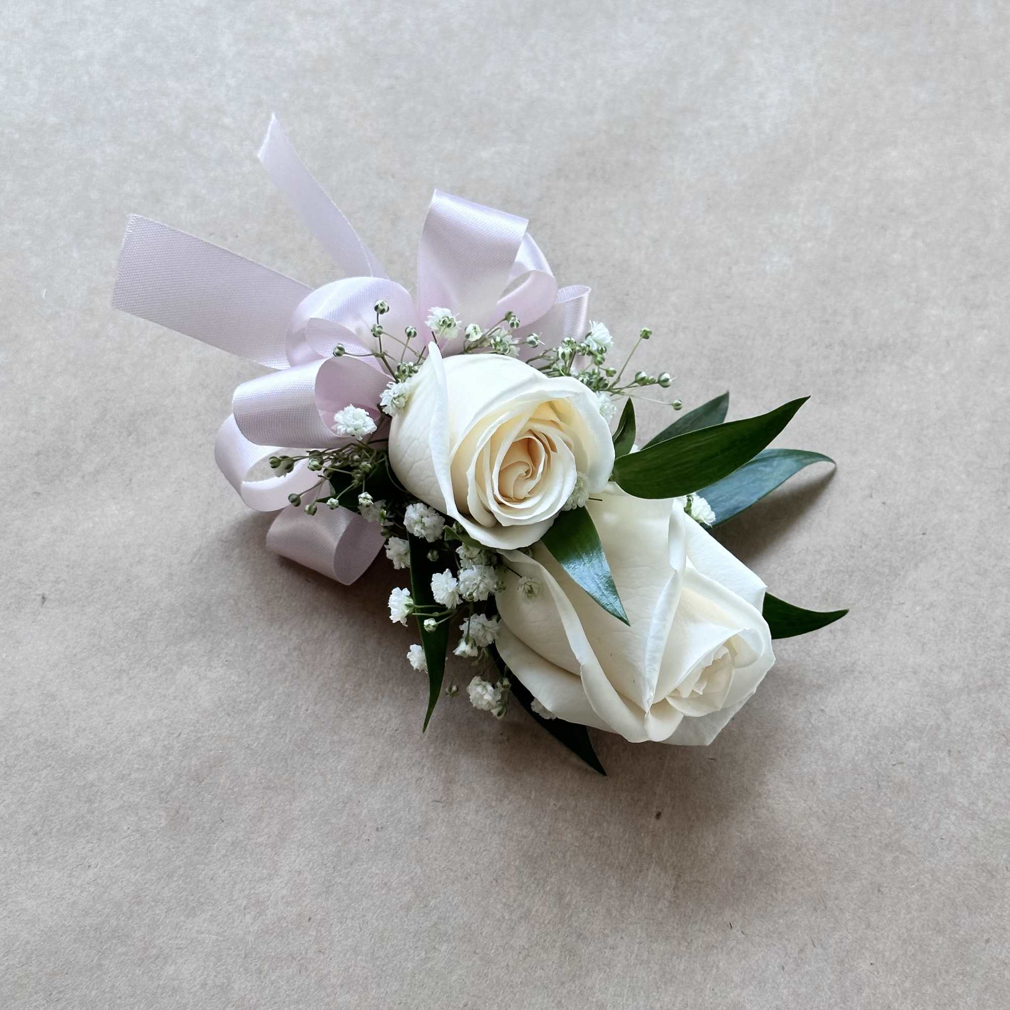 Corsage with two ivory roses, baby’s breath, and a pale pink ribbon bow on a neutral background