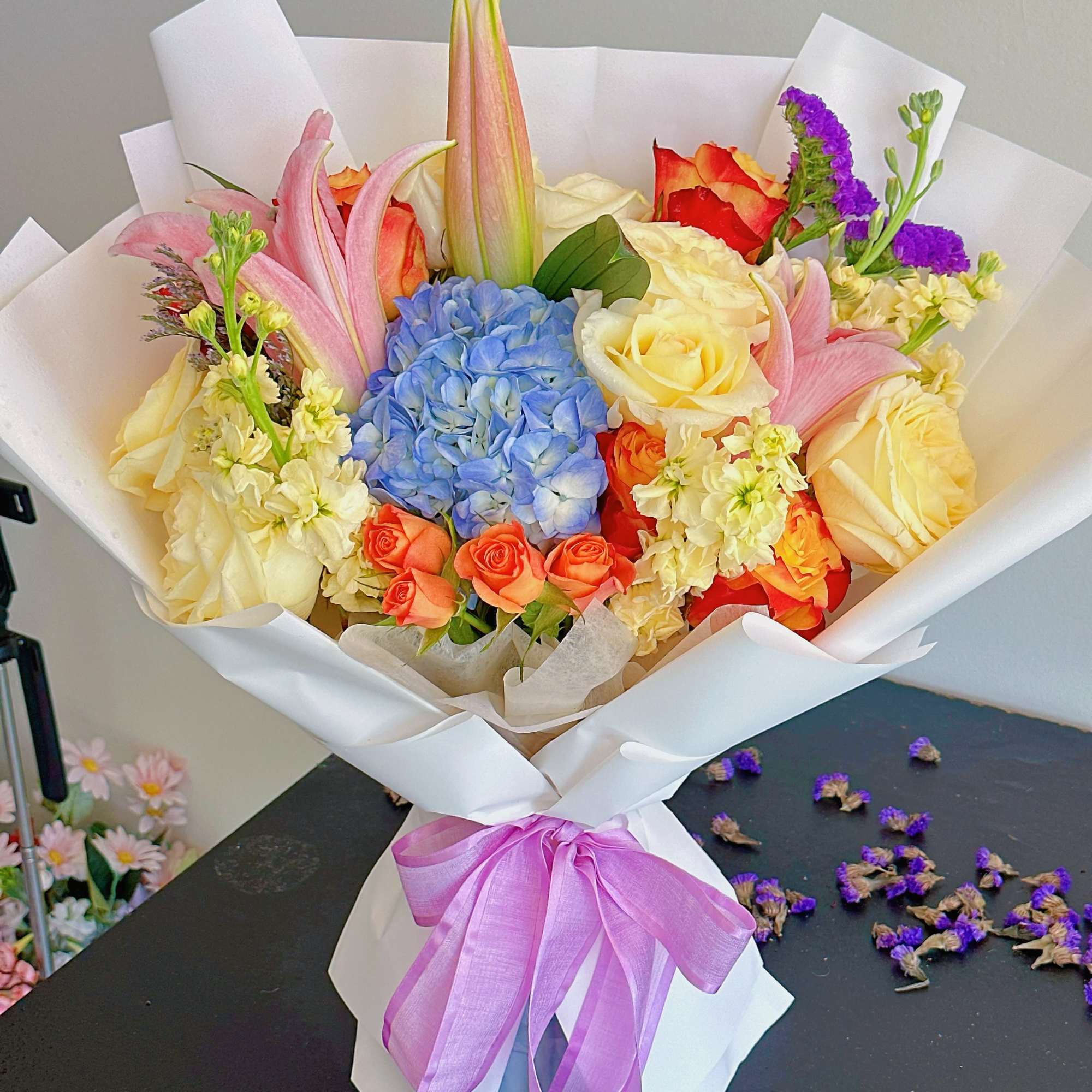 Bouquet of roses, lilies, and blue hydrangea wrapped in white paper with a purple ribbon