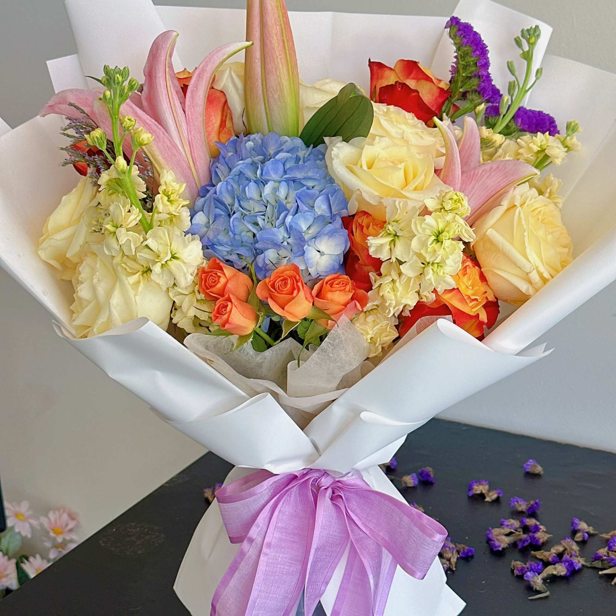Mixed bouquet of roses, hydrangea, and lilies wrapped in white paper with a purple ribbon