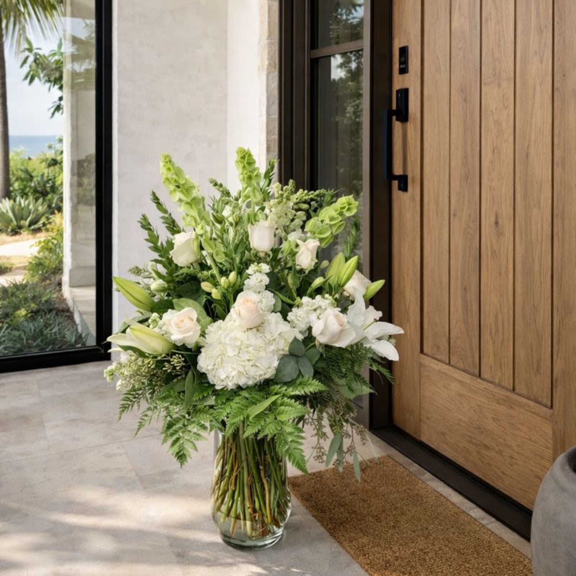 Tall white floral arrangement in a glass vase by a wooden door