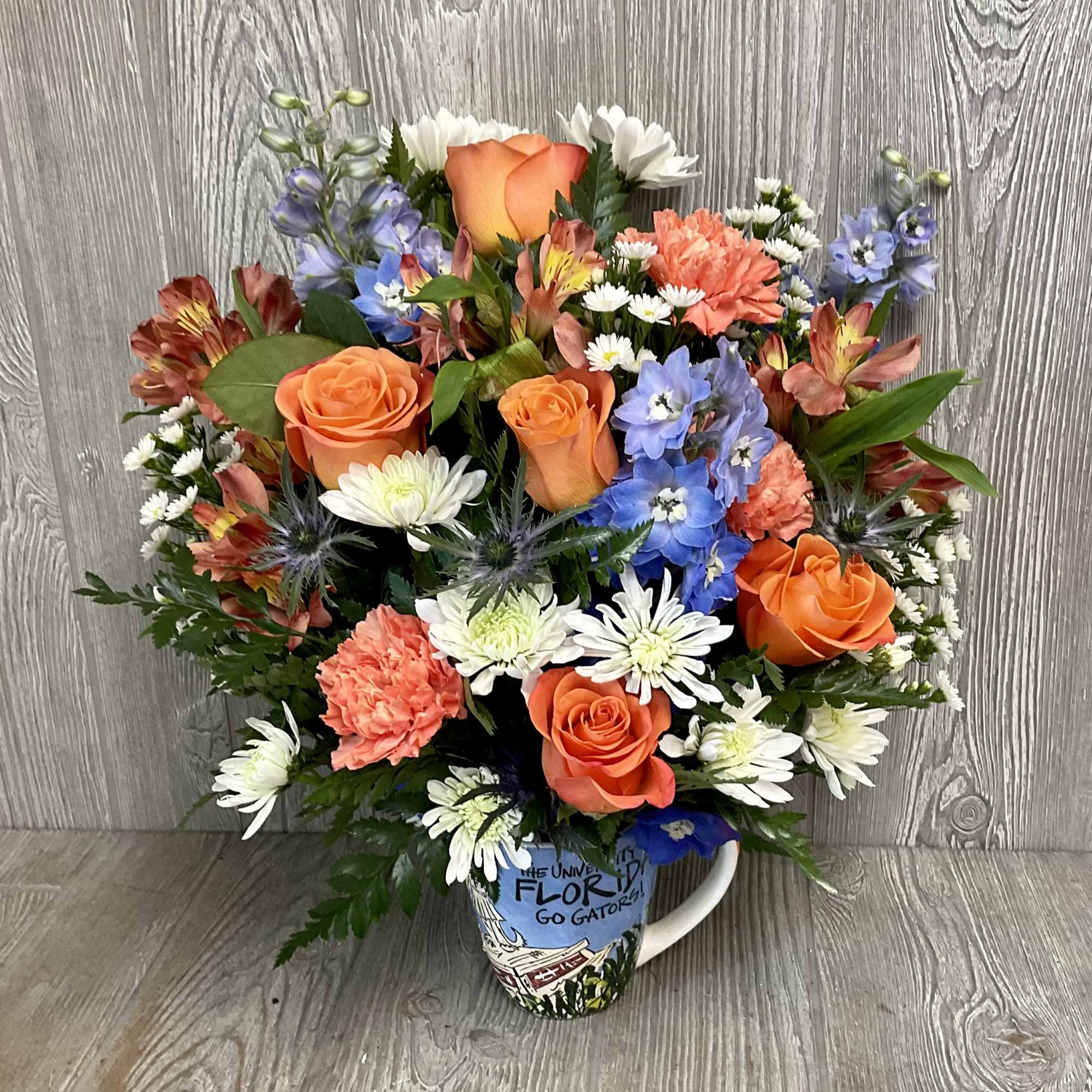 Mixed bouquet of orange roses, white daisies, and blue flowers in a printed mug