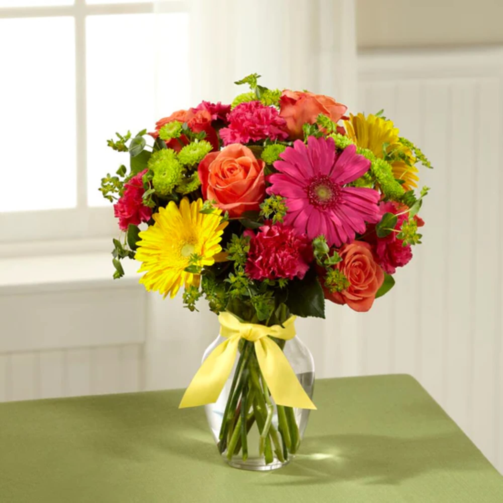 Mixed bouquet of roses, gerbera daisies, and carnations in a glass vase with a yellow ribbon