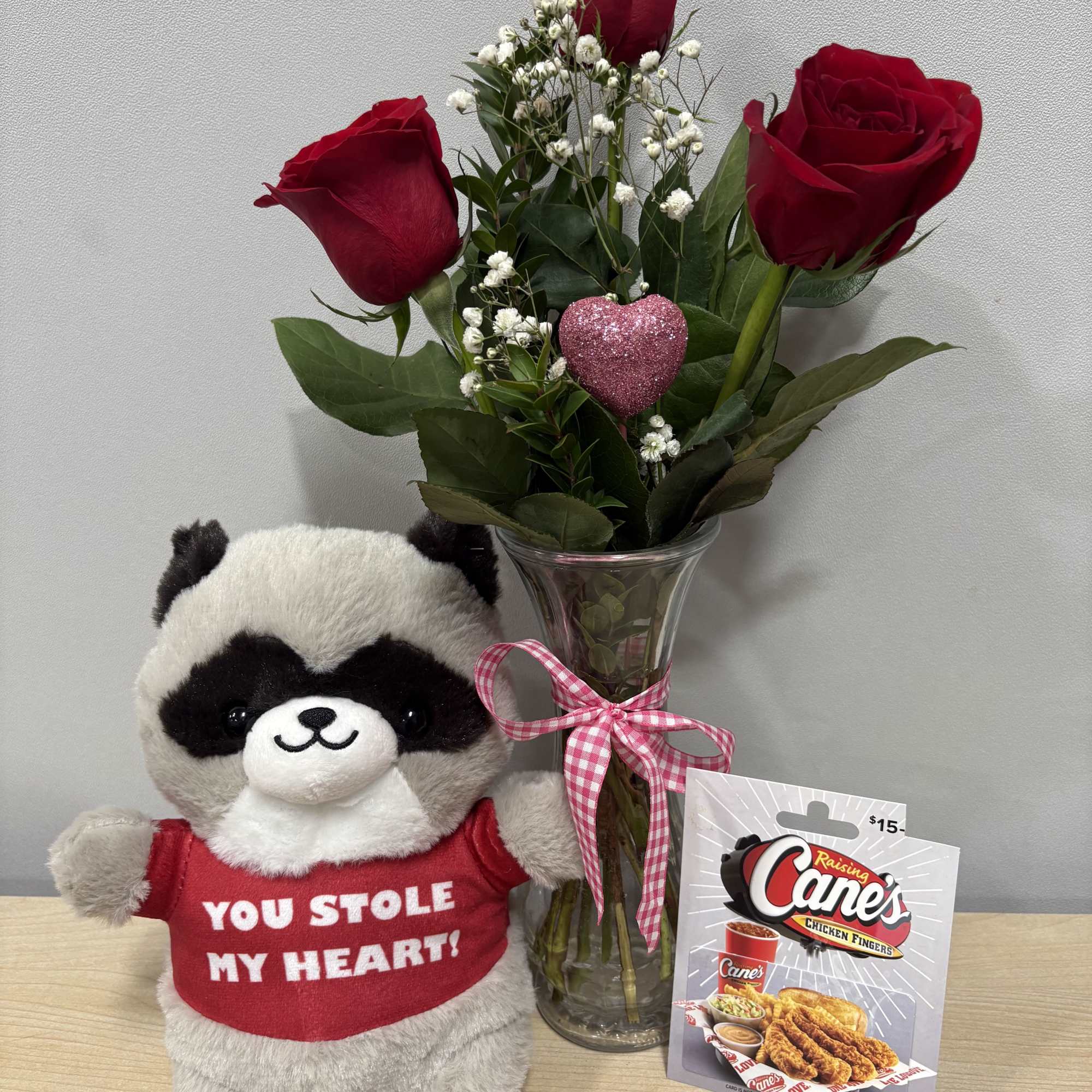 Three red roses in a glass vase beside a plush raccoon and gift card