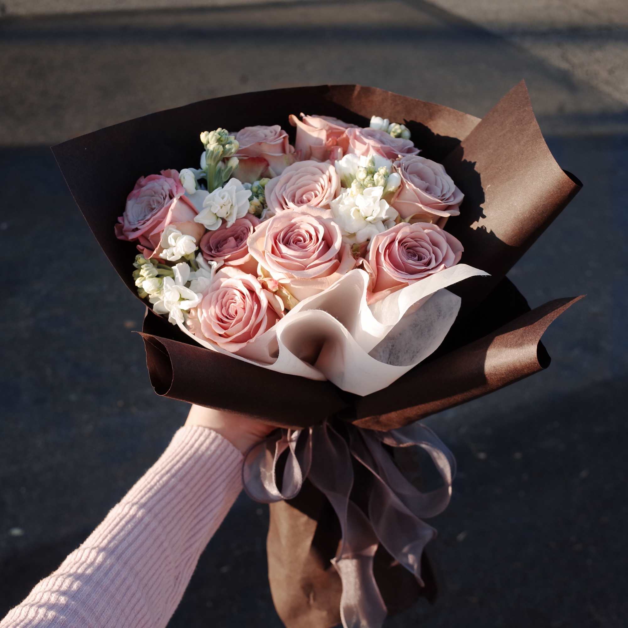 Handheld bouquet of pink roses and white filler flowers wrapped in dark paper