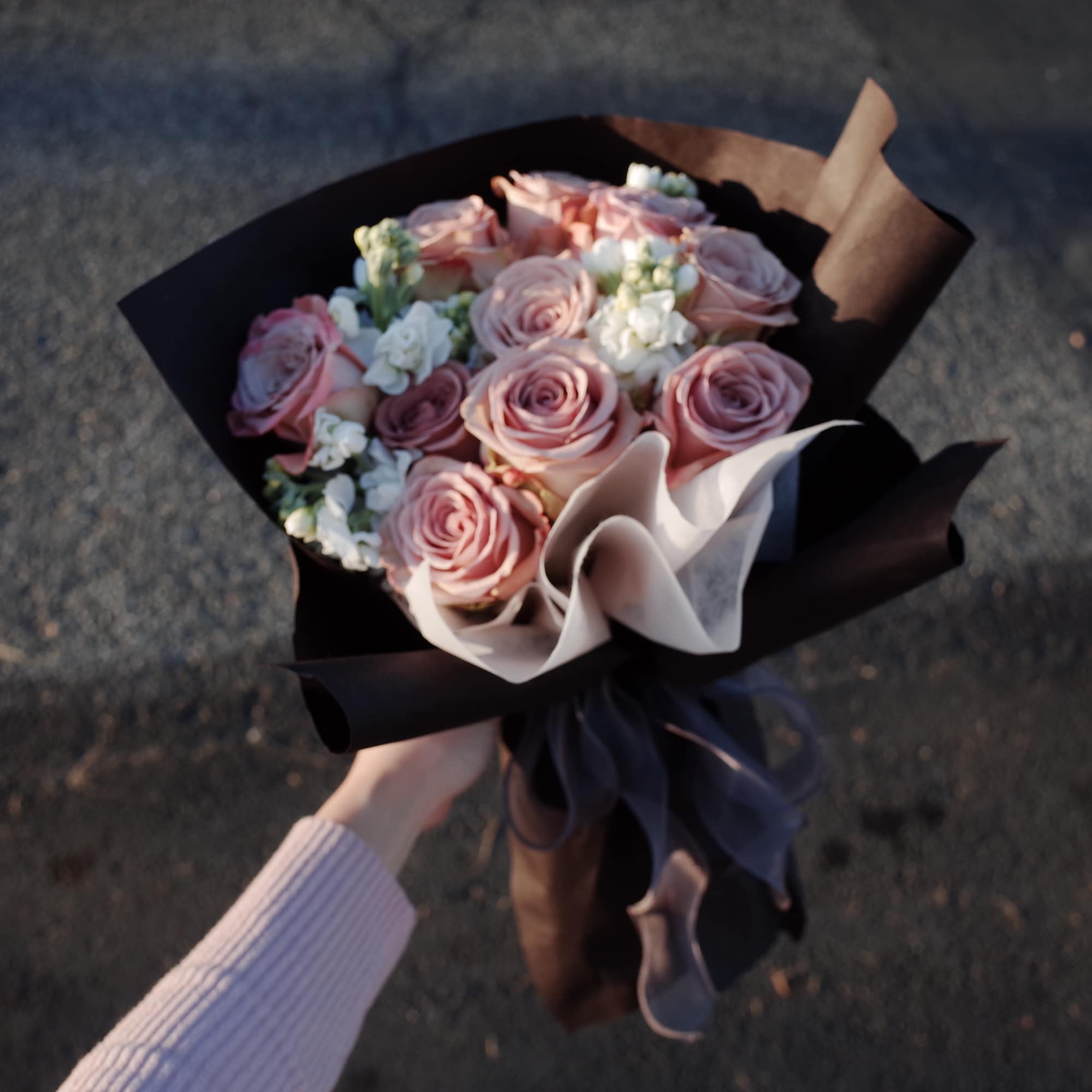 Handheld bouquet of pink roses with white filler flowers wrapped in dark paper